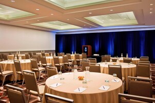 Un salón de conferencias con mesas redondas, sillas color beige y un escenario con un telón de fondo azul.