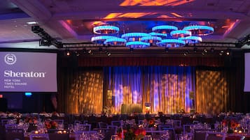 Een feestzaal met ronde tafels gedekt voor een formeel evenement, een podium met een groot scherm en een kroonluchter.