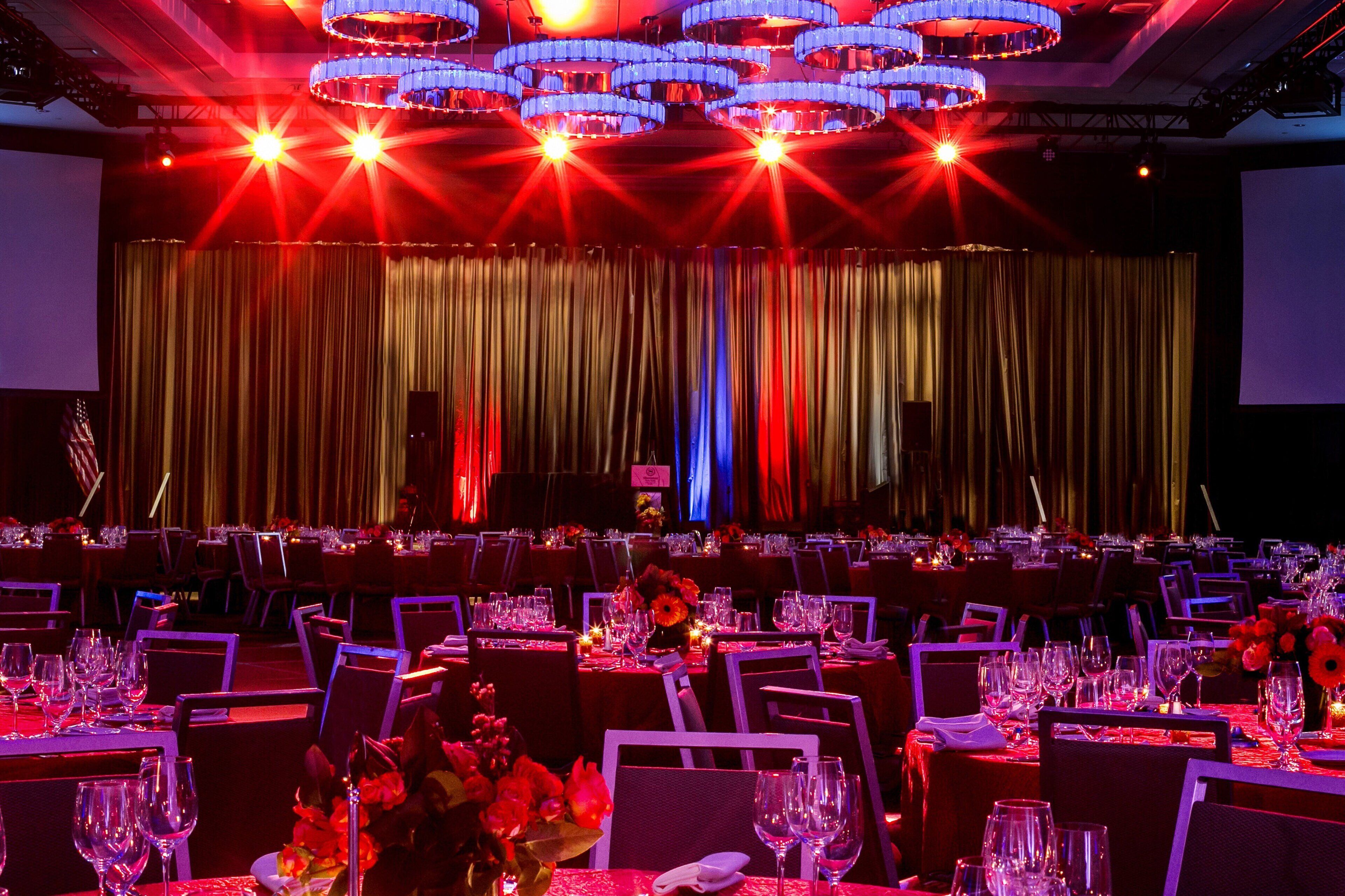 A banquet hall with tables set for a formal event, featuring a stage with curtains and overhead lighting.