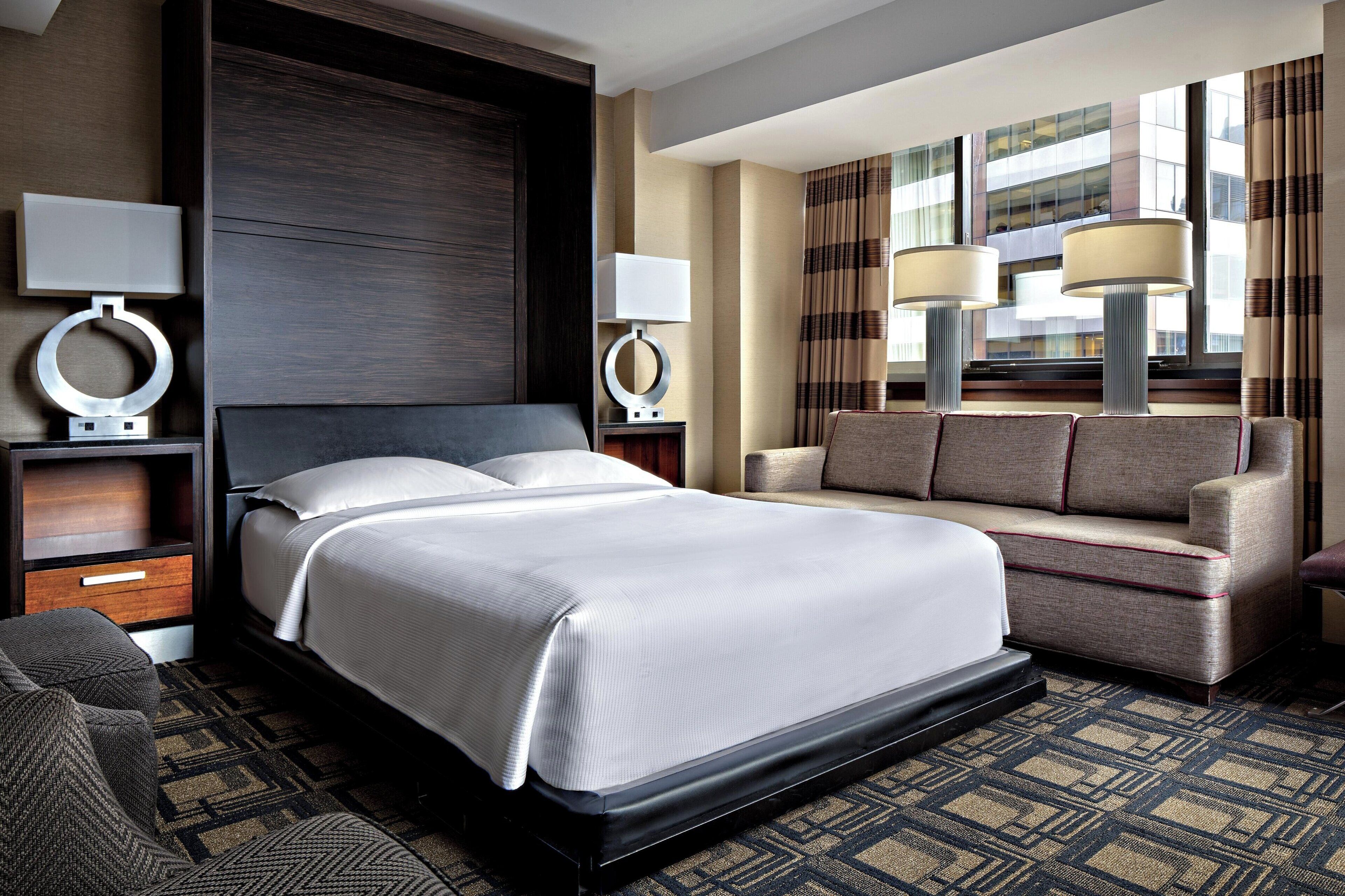 A hotel room with a large bed, a sofa, a desk with a lamp, and a view of a building through the window.