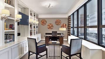 A modern hotel room with a large window, a desk with two chairs, a bookshelf, and decorative wall art.