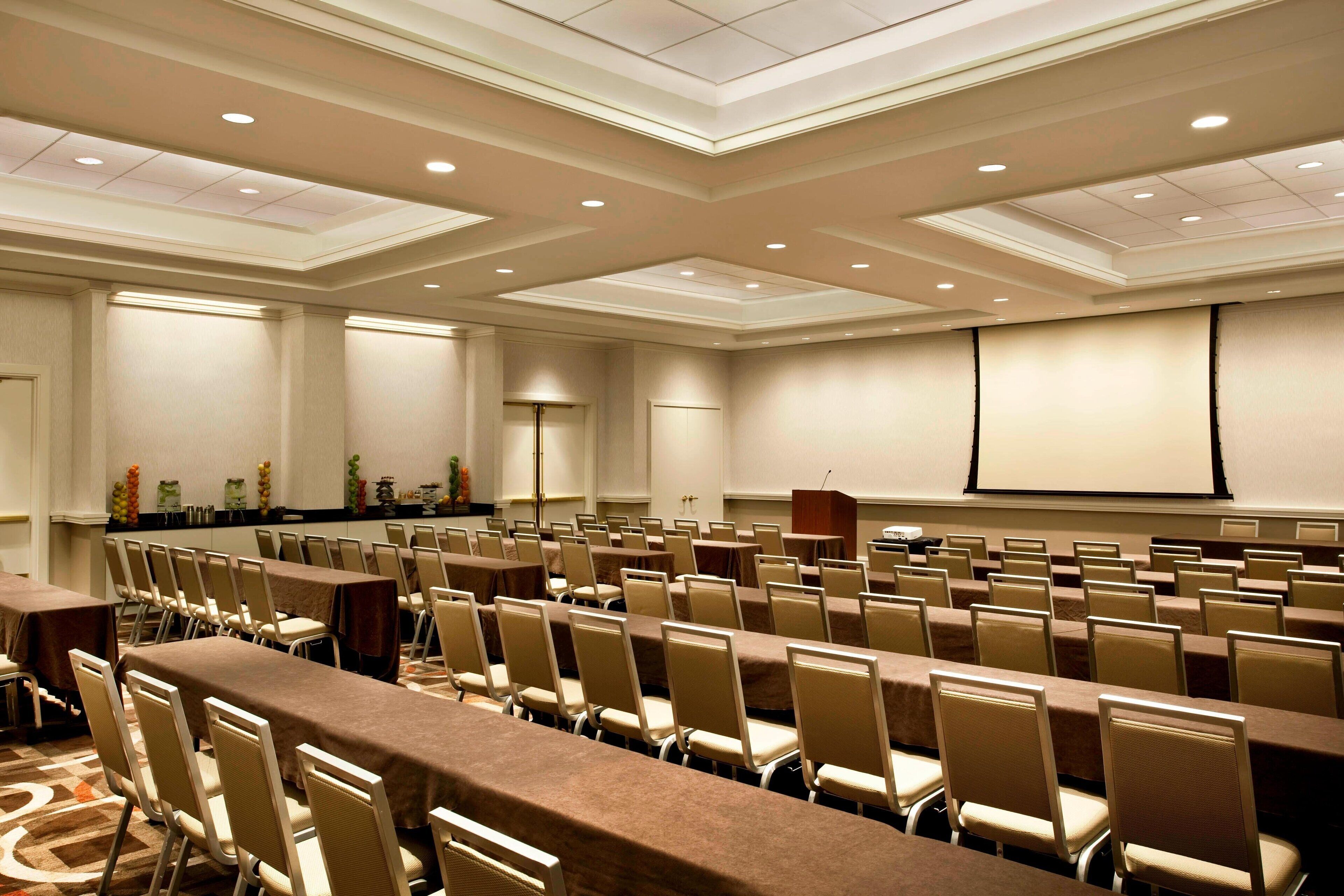 A conference room with rows of tables and chairs, a podium, and a projector screen.