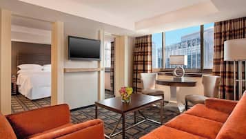 Quarto de hotel com cama, televisão, uma pequena mesa com um vaso, cadeiras laranja e vista para a cidade.