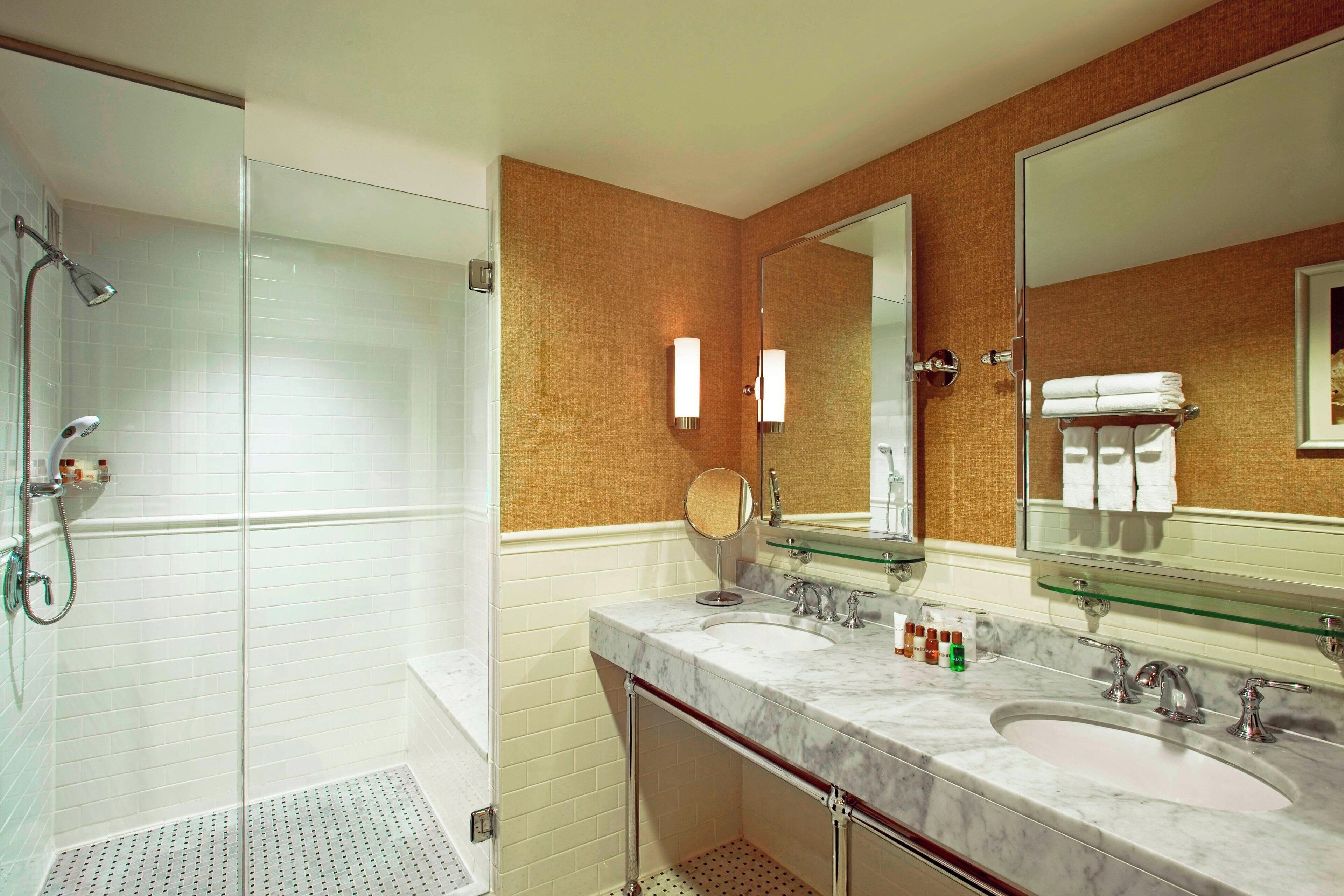 Un baño con ducha envuelta en vidrio, dos lavamanos con espejos y un perchero para toallas.