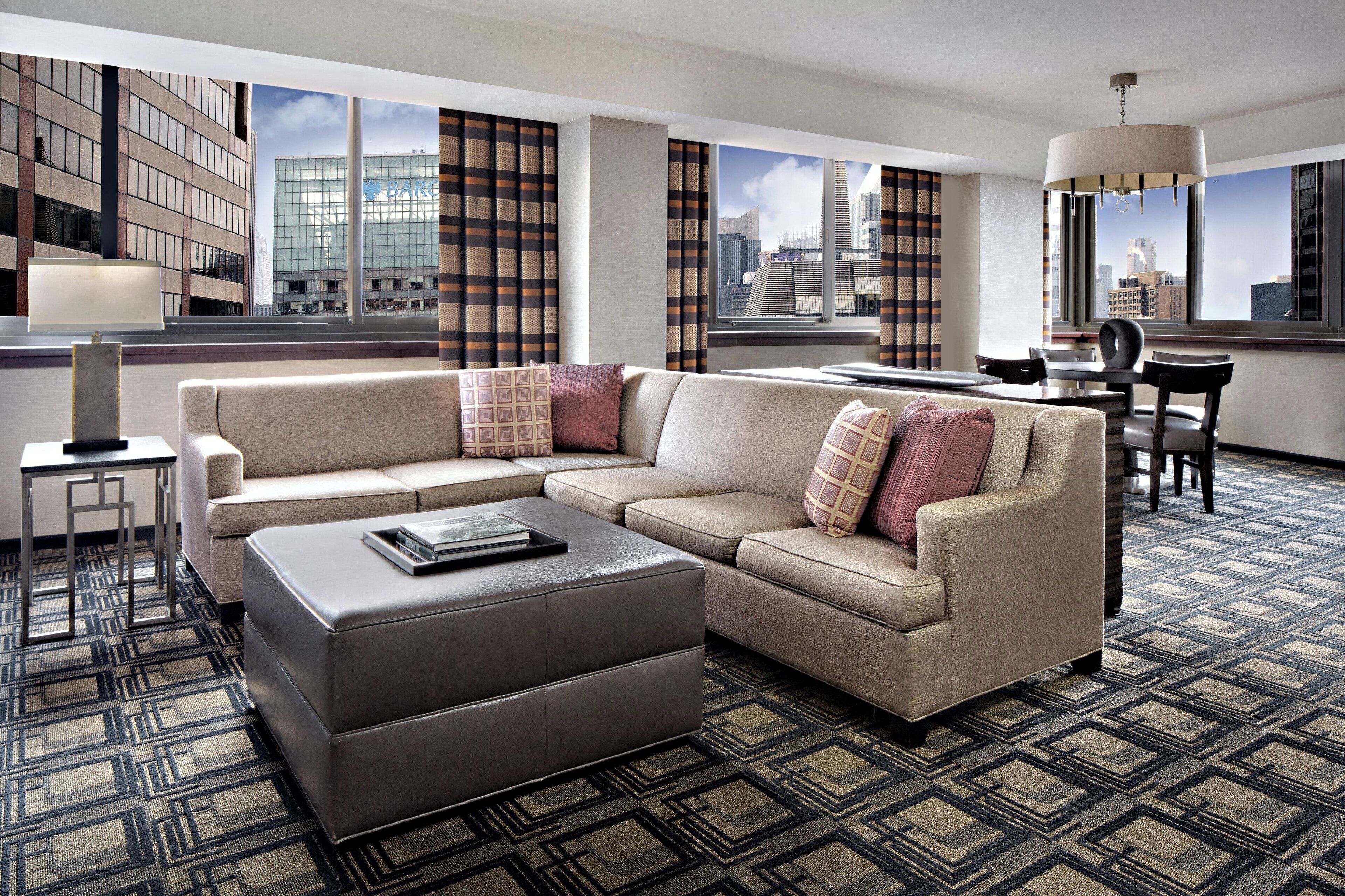 Ein modernes Hotelzimmer mit Ecksofa, einem kleinen Tisch und Blick auf die Stadt durch große Fenster.