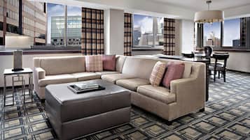 A modern hotel room with a sectional sofa, a small table, and a view of the cityscape through large windows.