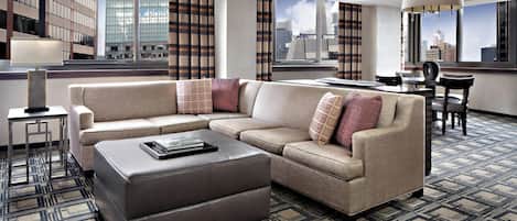 Ein modernes Hotelzimmer mit Ecksofa, einem kleinen Tisch und Blick auf die Stadt durch große Fenster.