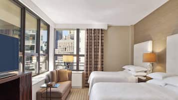 Ein Hotelzimmer mit zwei Betten, einem Sofa, einem Fernseher und einem Fenster mit Blick auf die Stadt.