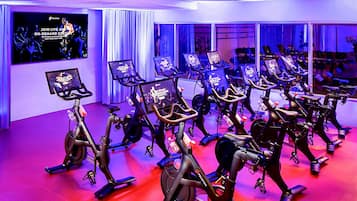Un estudio de fitness con bicicletas estáticas dispuestas en filas, una pantalla grande mostrando una clase bajo demanda y un muro espejo que refleja las bicicletas.