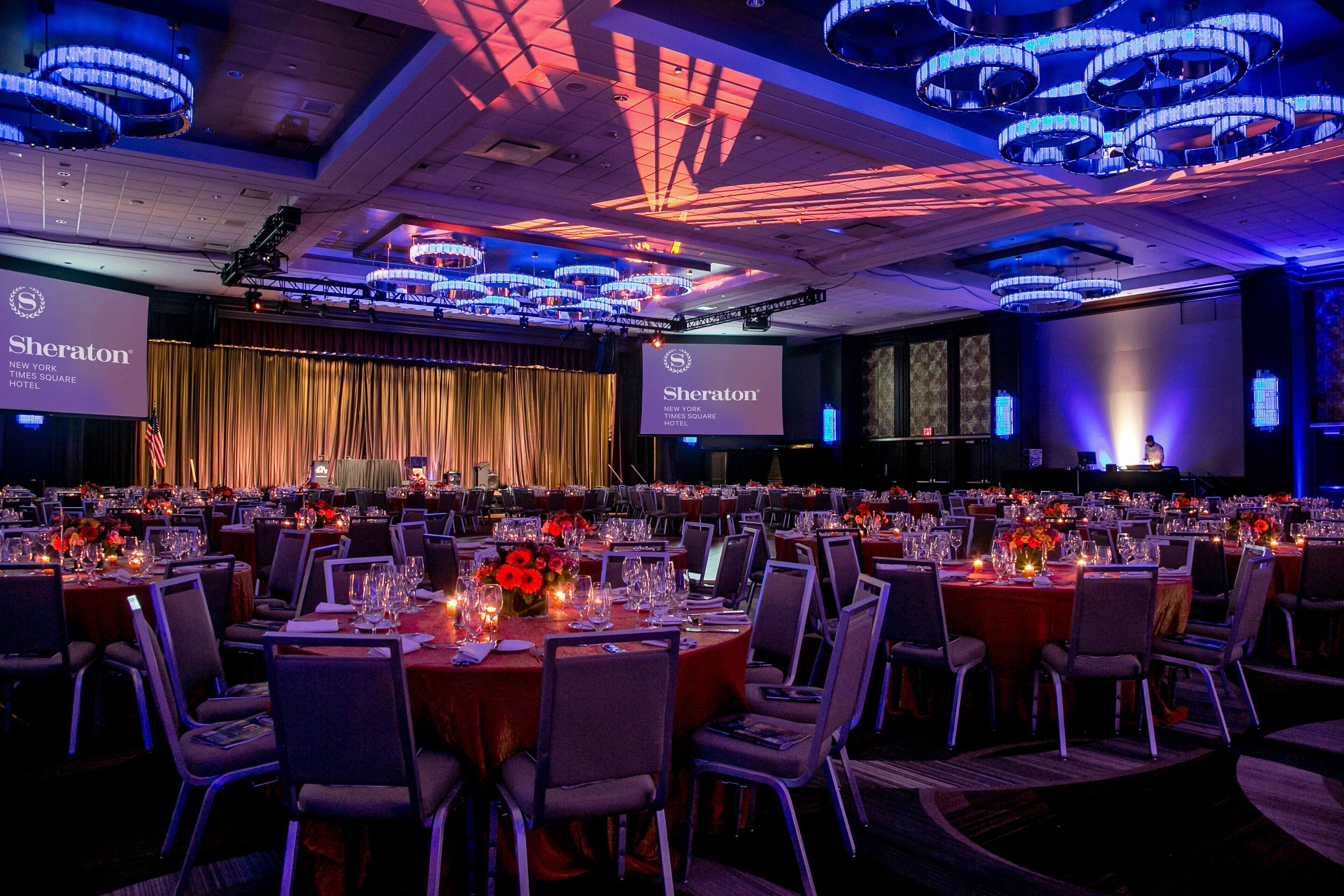 A Sheraton hotel ballroom with rows of round tables set for dining, a stage with curtains, and a large screen displaying the hotel's name.