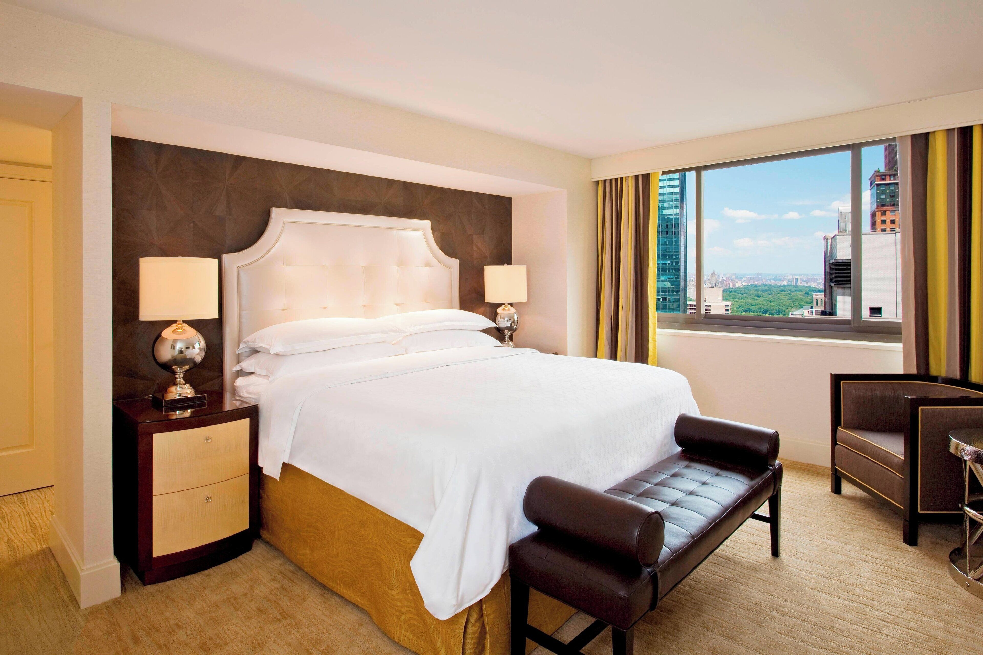 A hotel room with a large bed, two bedside tables with lamps, a bench, a desk, and a large window with a city view.