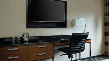 A hotel room with a flat-screen TV mounted on the wall, a wooden desk with a black chair, and a small table with a kettle and glasses.