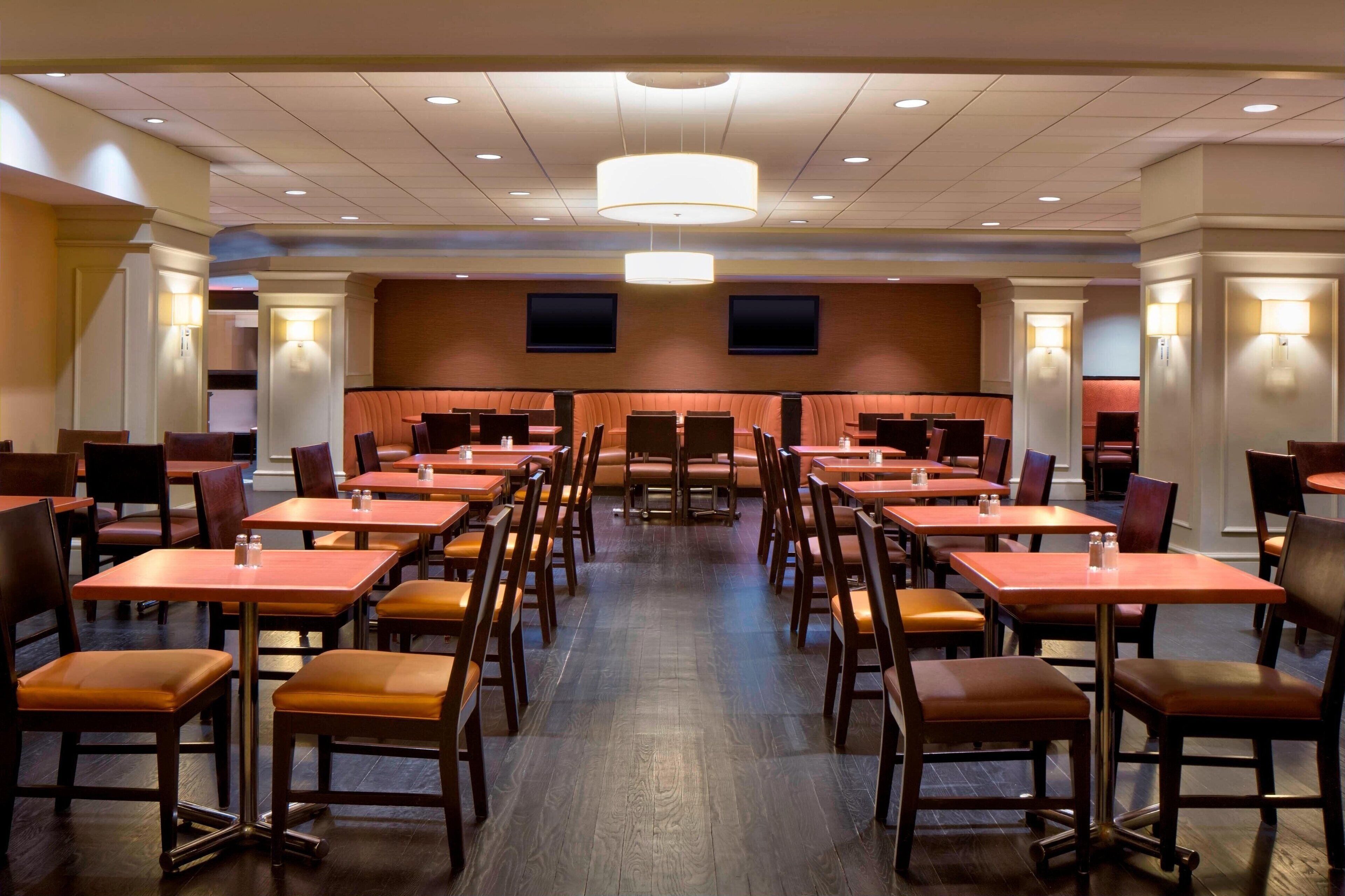 A spacious hotel dining area with multiple tables and chairs, recessed ceiling lighting, and a central seating area.