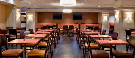 A spacious hotel dining area with multiple tables and chairs, recessed ceiling lighting, and a central seating area.