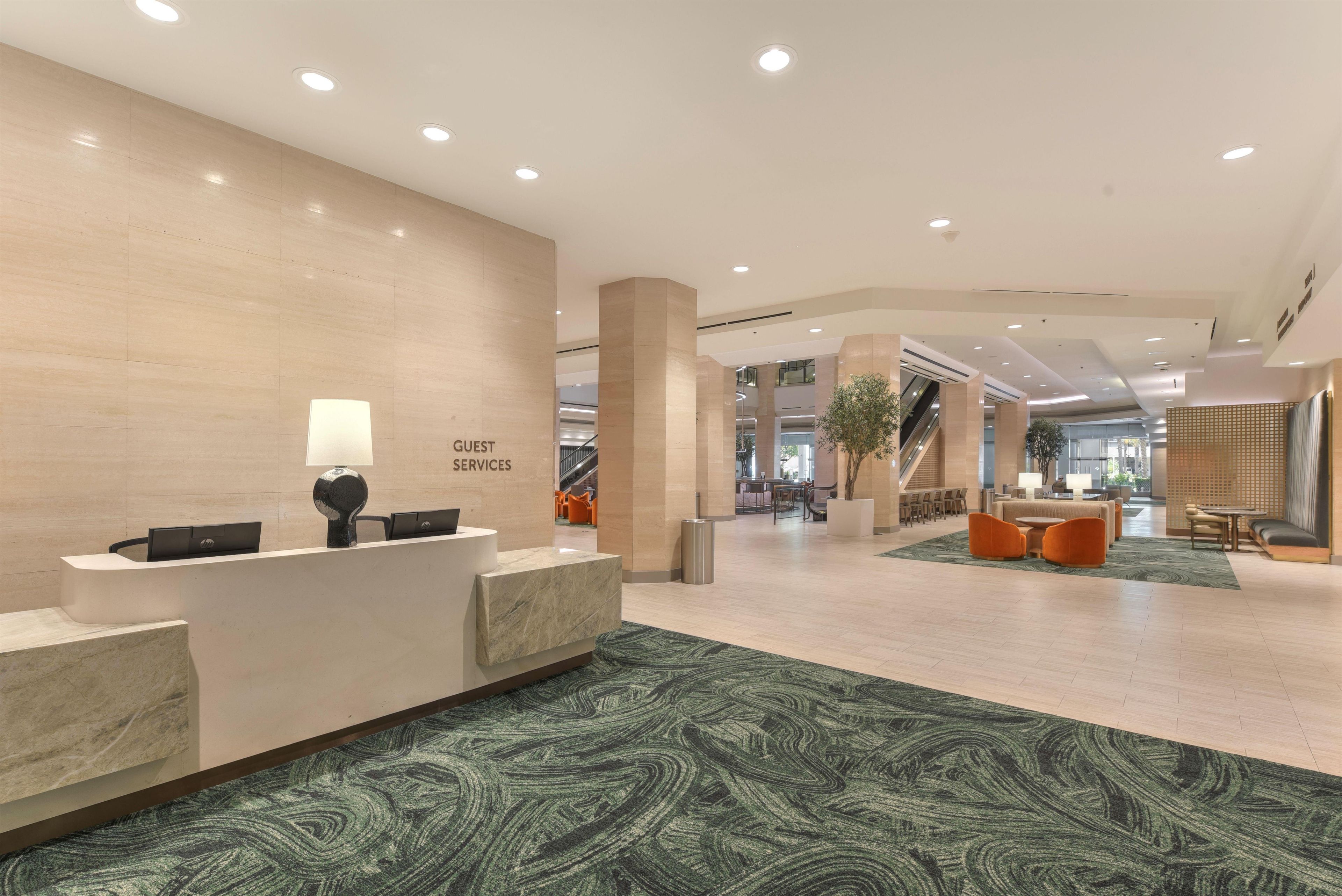 Zona de recepción de hotel con mostrador de mármol, cartel de servicios para huéspedes y alfombra estampada verde.