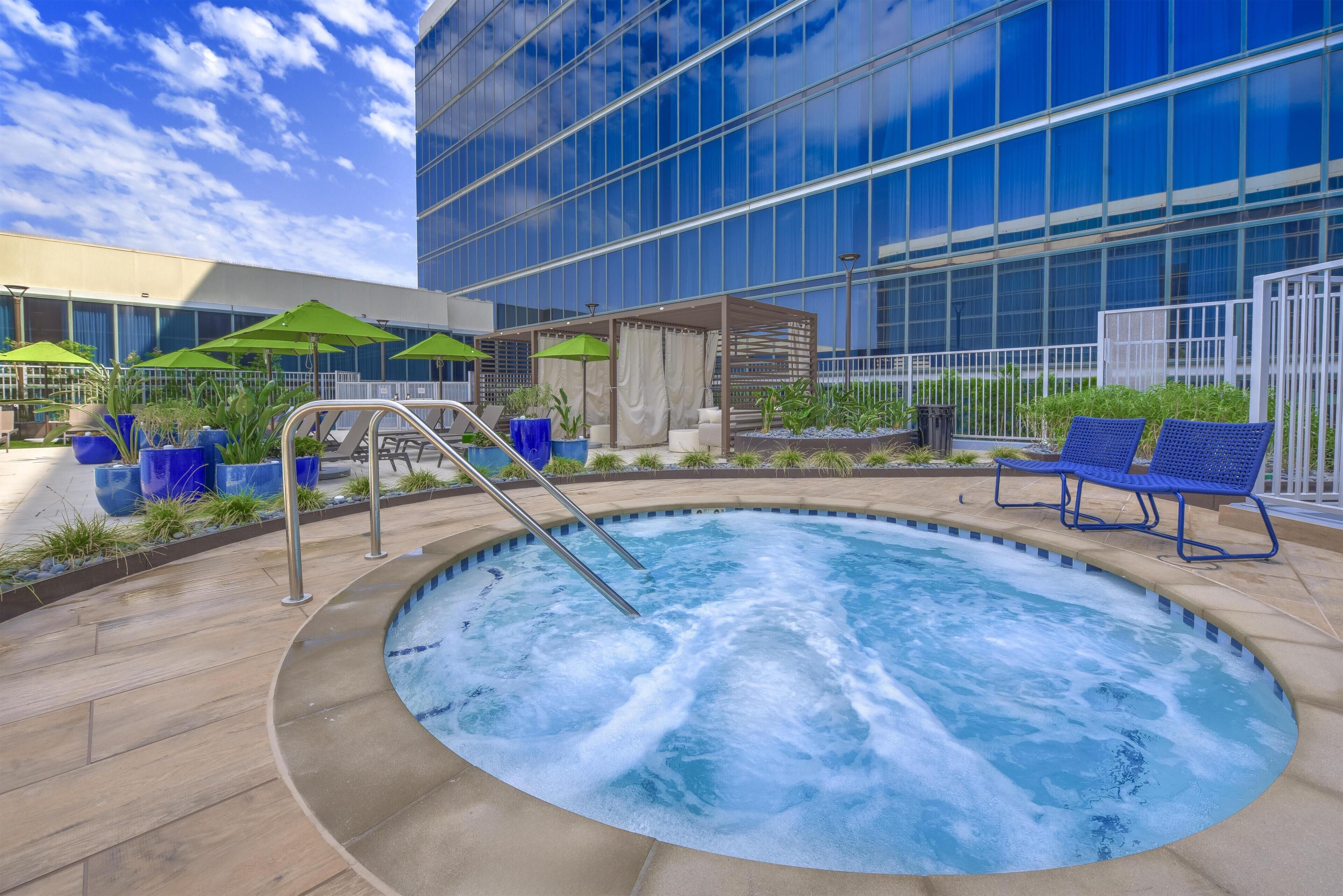 A hot tub with a metal ladder, surrounded by blue chairs and green umbrellas, in front of a modern building with large windows.