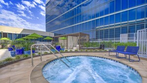 A hot tub with a metal ladder, surrounded by blue chairs and green umbrellas, in front of a modern building with large windows.