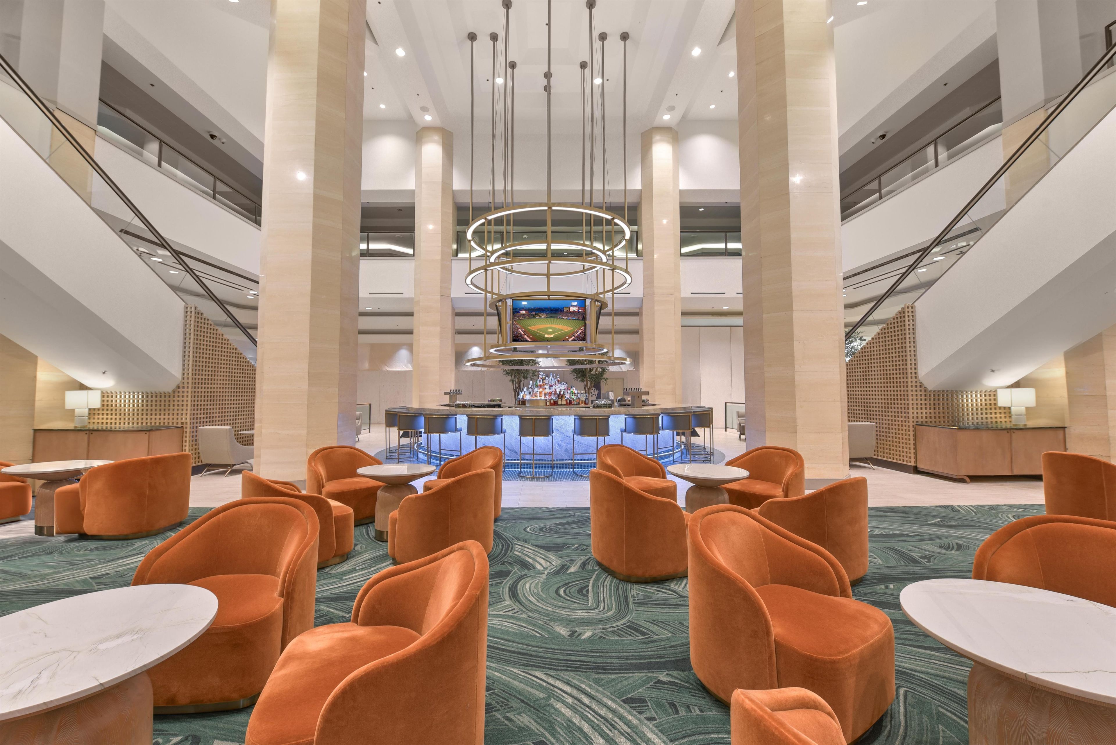 A spacious lobby with a central bar area, multiple seating options, and a large screen displaying a baseball game.