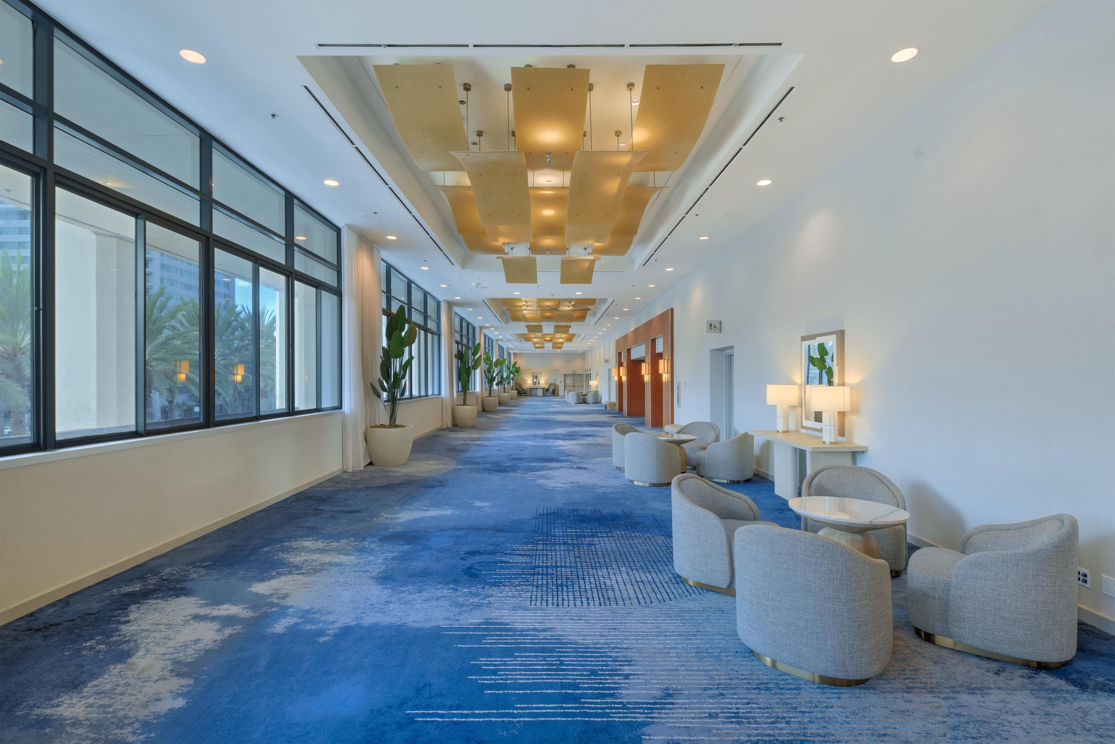 A hotel corridor with a blue carpet, seating area with chairs, and large windows on one side.