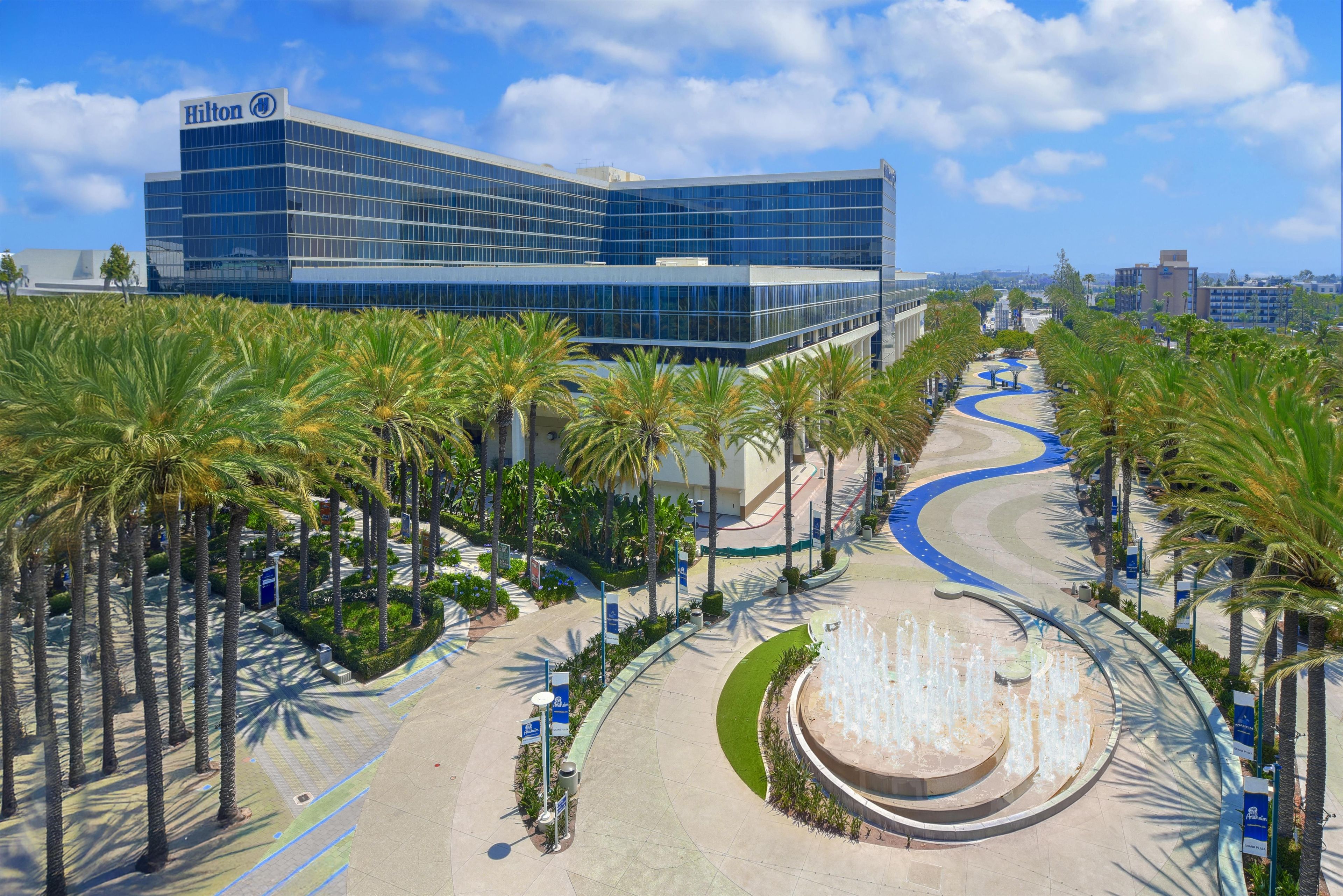 A modern hotel with a glass facade, palm trees, and a central water feature.