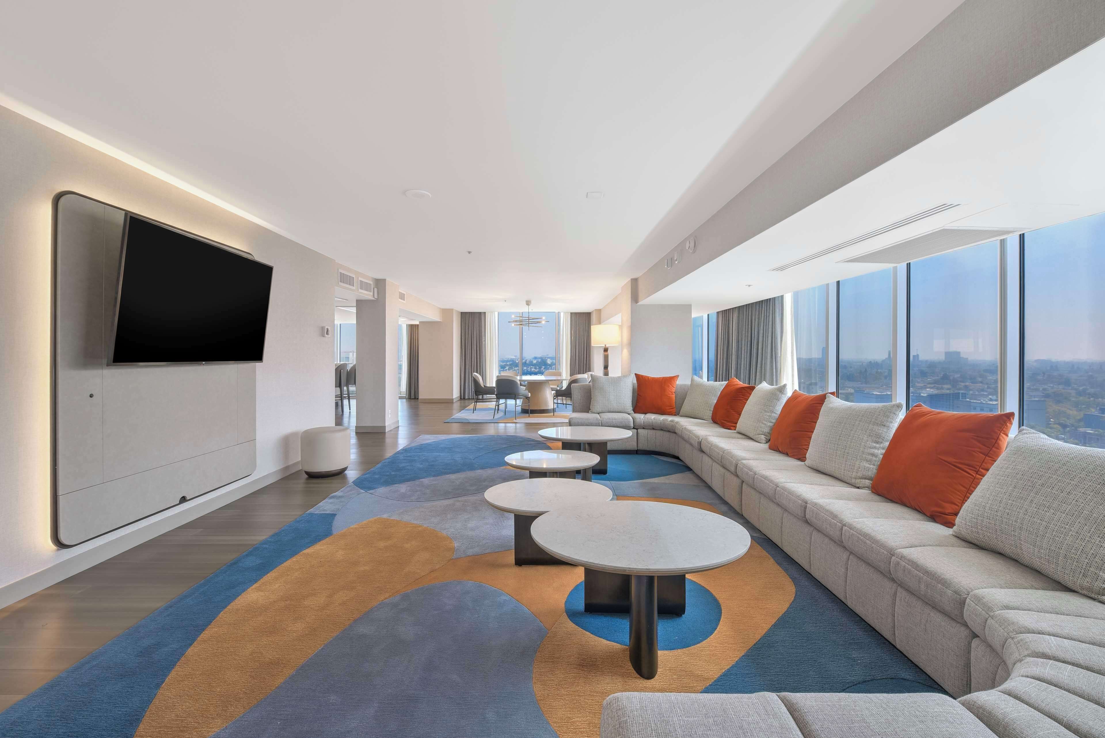 A modern hotel room with a large sofa, round tables, a flat-screen TV, and a city view.