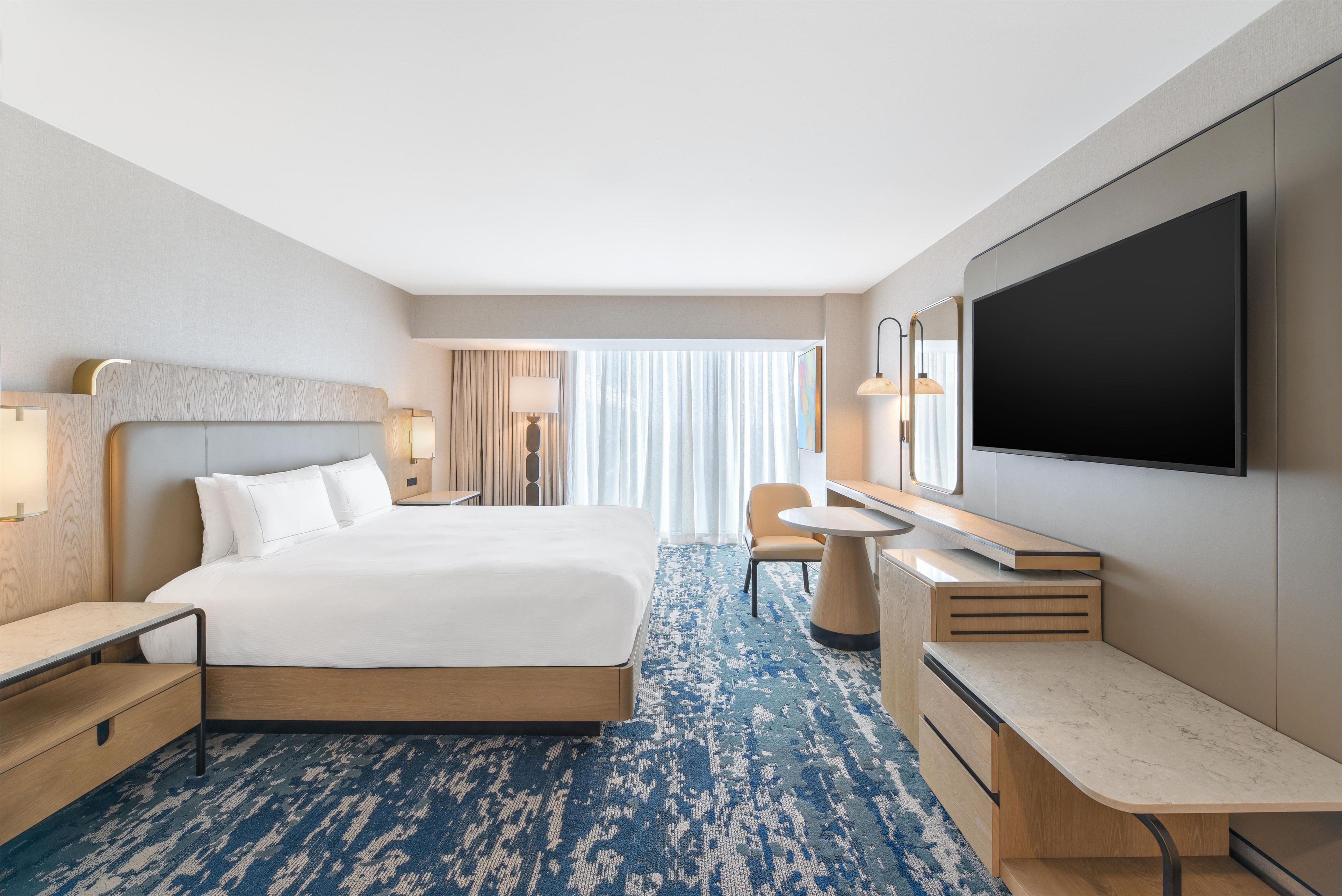 A hotel room with a bed, bedside tables, a desk, a TV, and a large window with curtains.