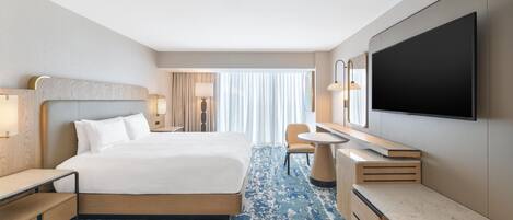 A hotel room with a bed, bedside tables, a desk, a TV, and a large window with curtains.