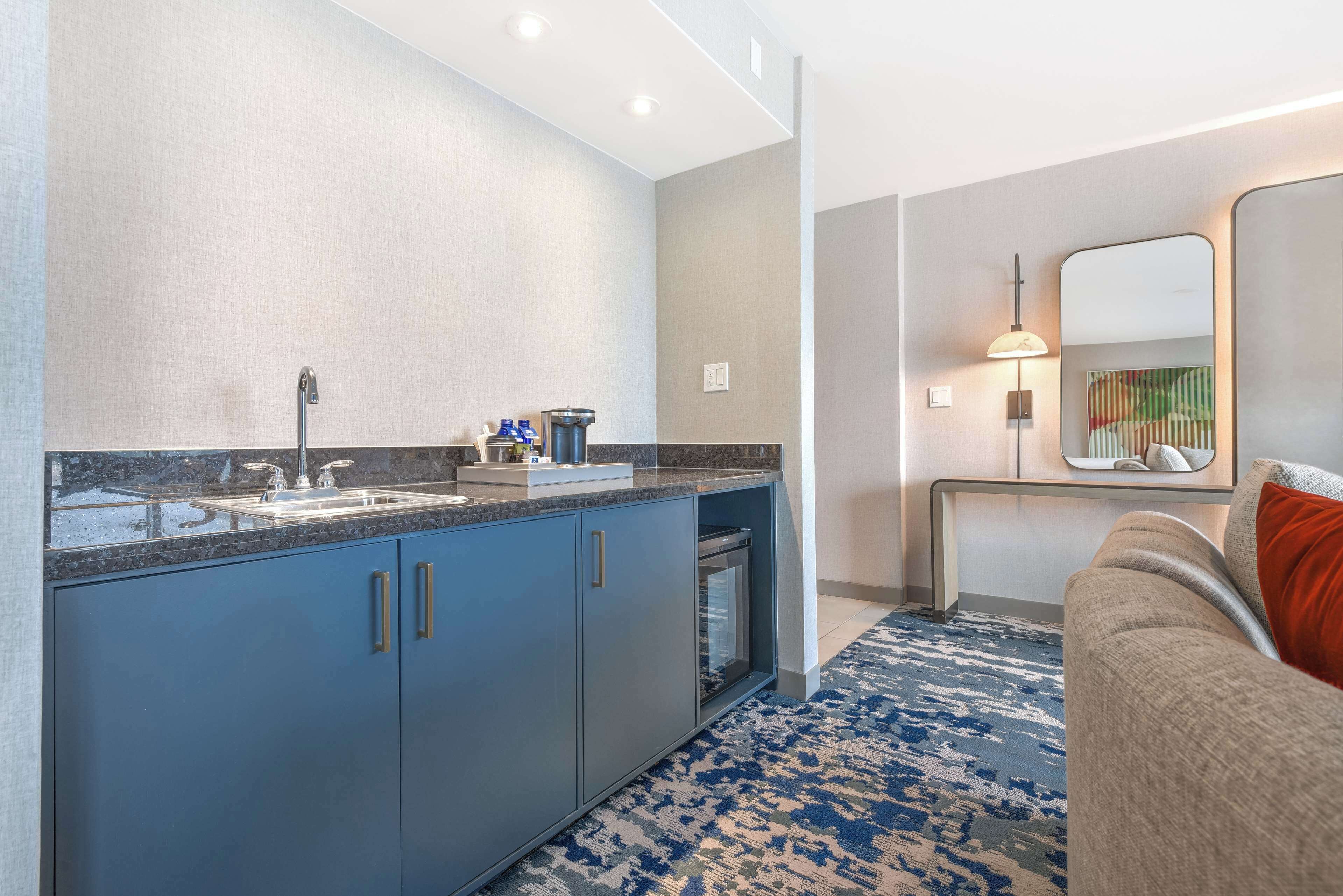 A modern kitchen with a sink, countertop, and cabinets. There is a coffee maker and a mirror above the counter. A patterned rug is on the floor, and a sofa is in the background.