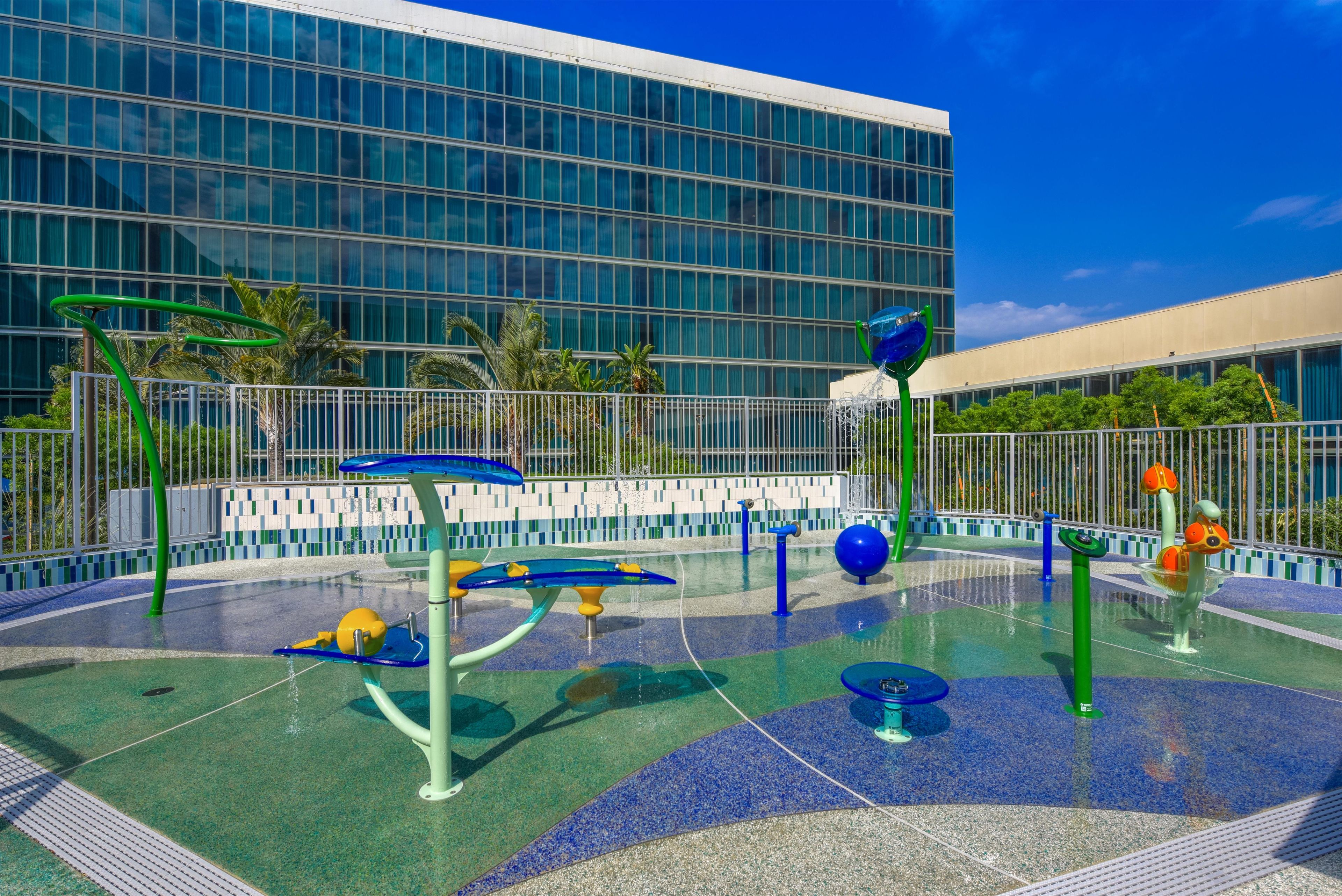 Zona de juegos al aire libre con diferentes fuentes de agua y un edificio moderno al fondo.
