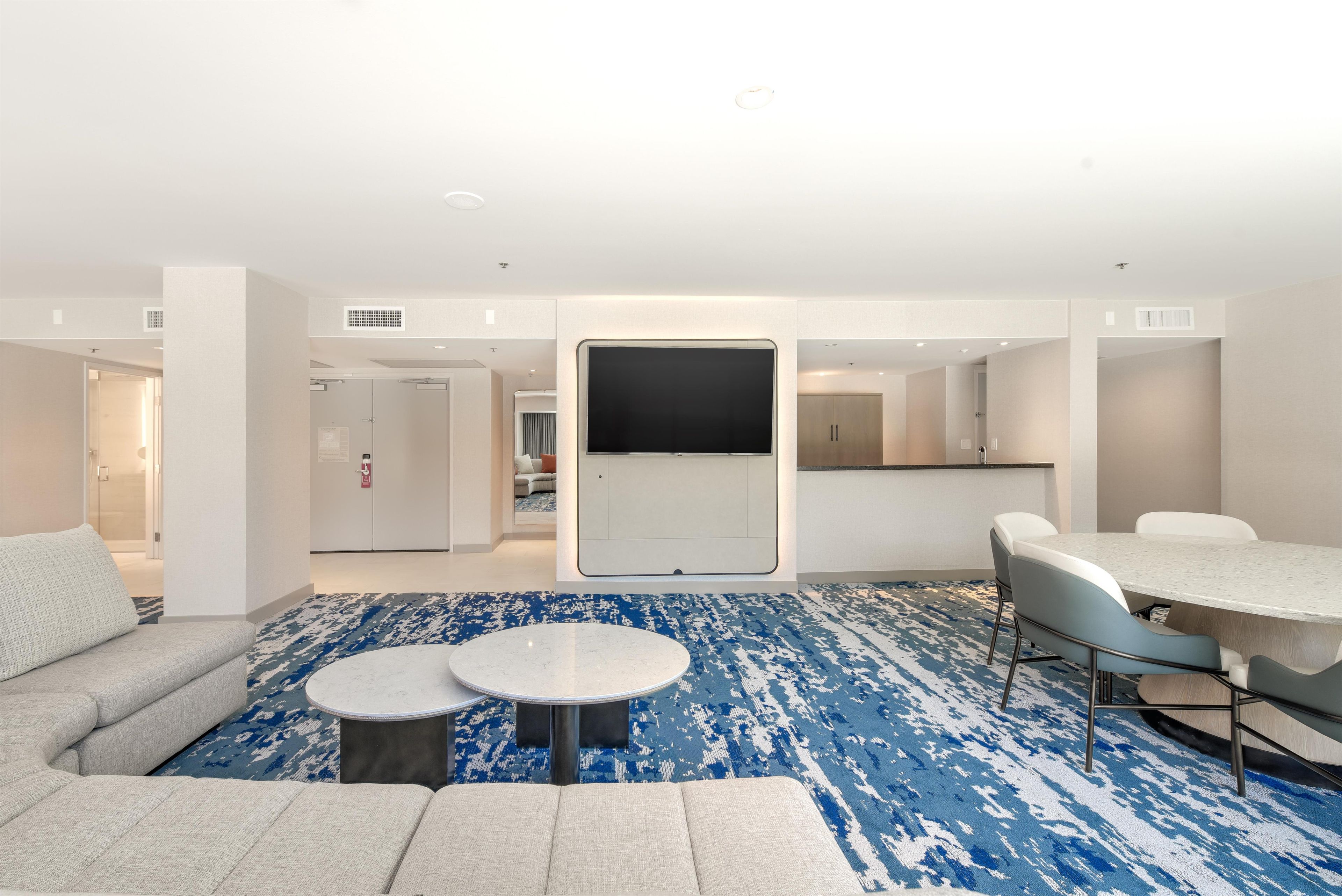 A modern hotel room with a sofa, two round tables, a dining area, and a large mirror.