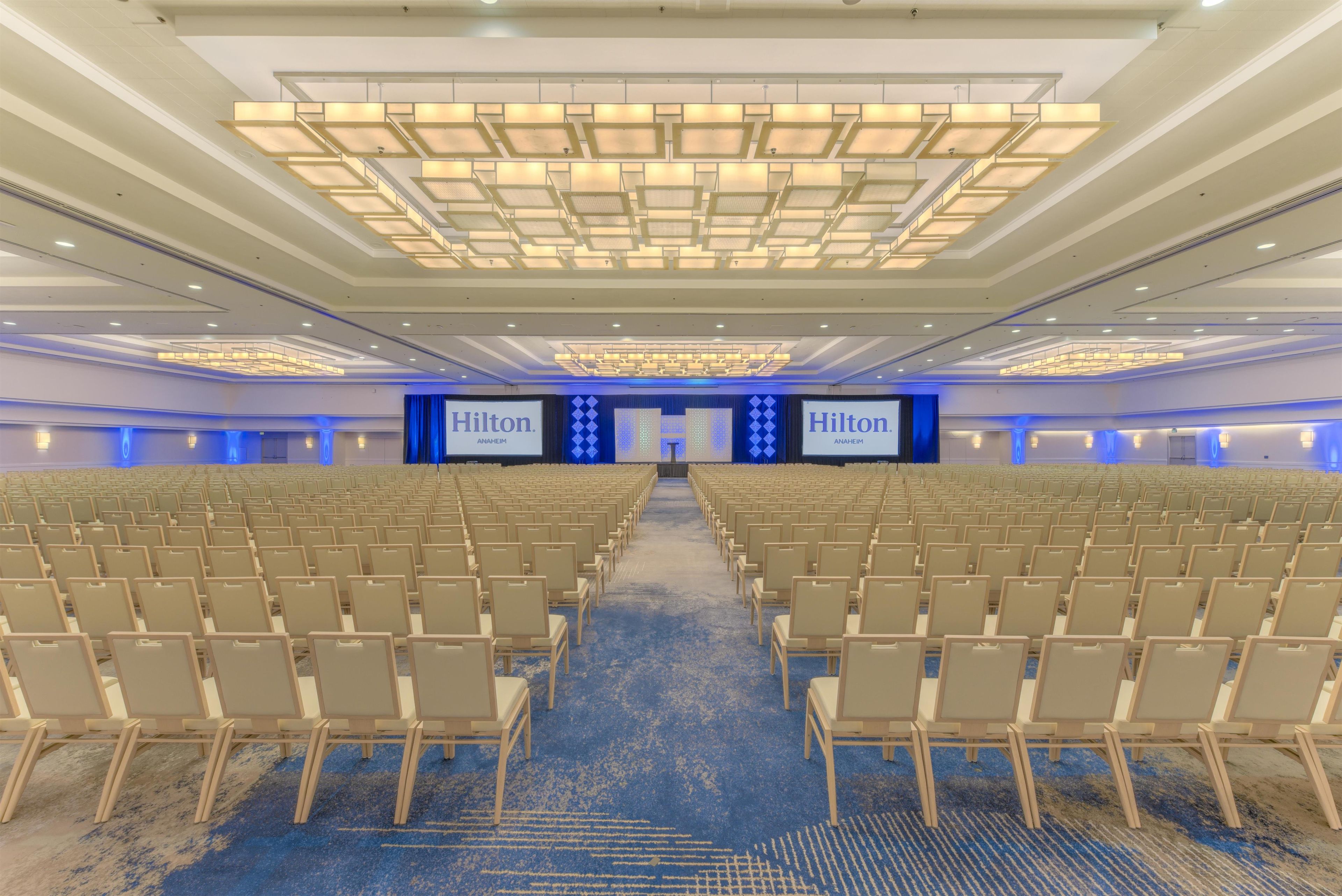 A large conference room with rows of chairs, a stage with a large screen, and a modern ceiling design.