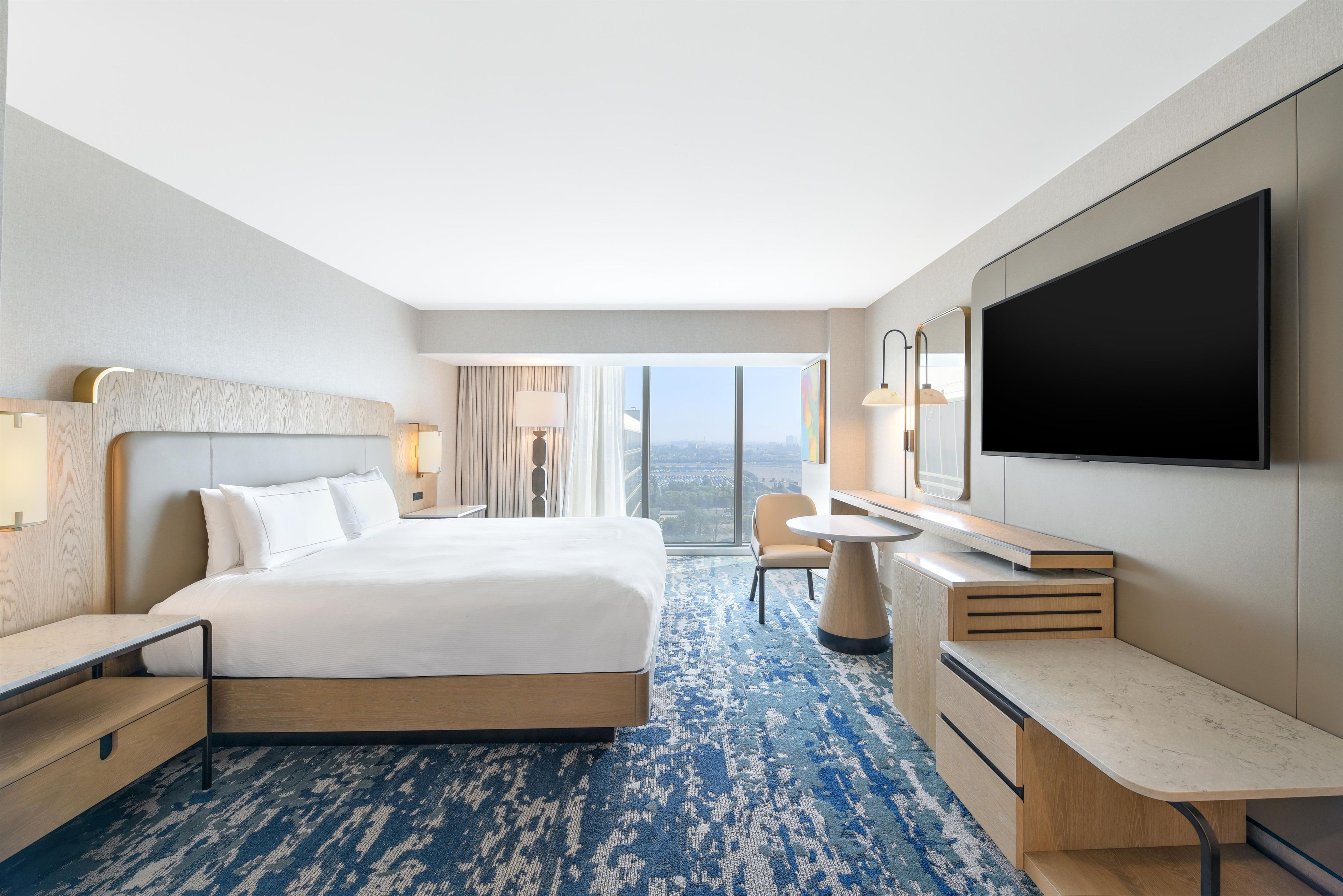 A hotel room with a bed, desk, chair, and a large TV on the wall.