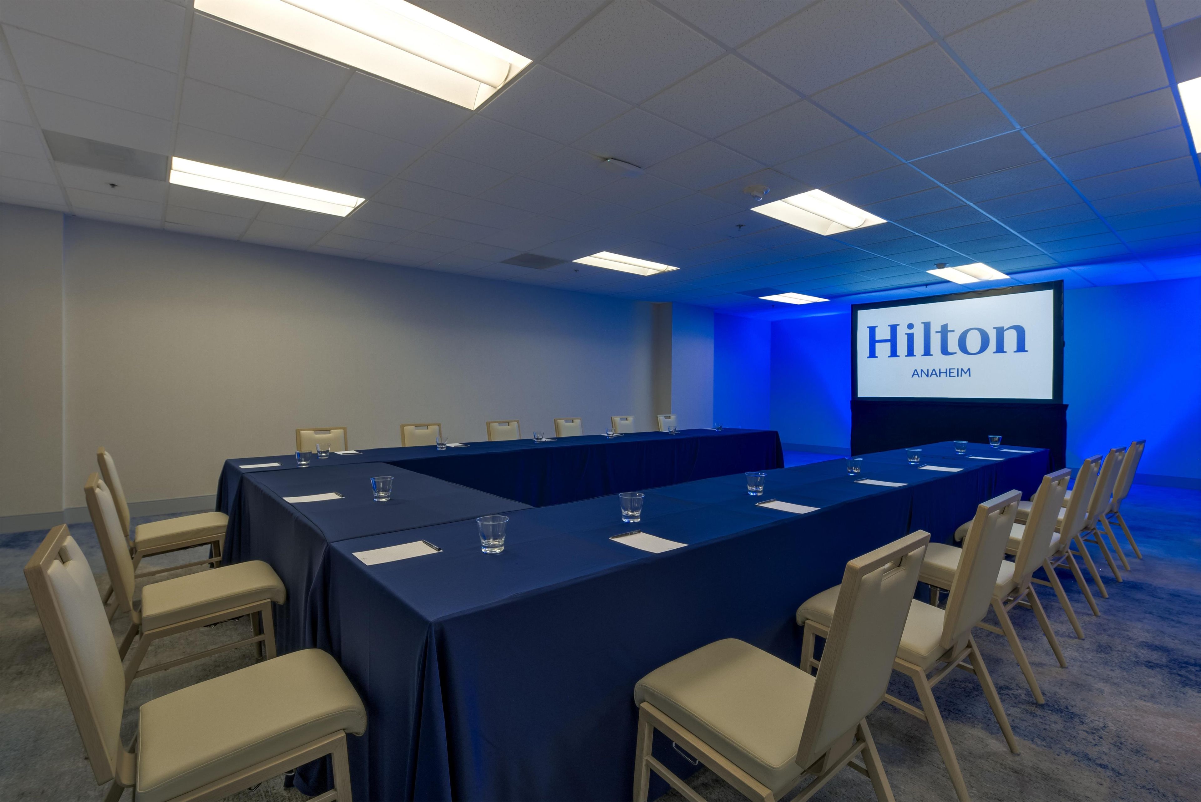 Un salón de conferencias con una larga mesa, sillas y una pantalla que muestra 'Hilton Anaheim'.