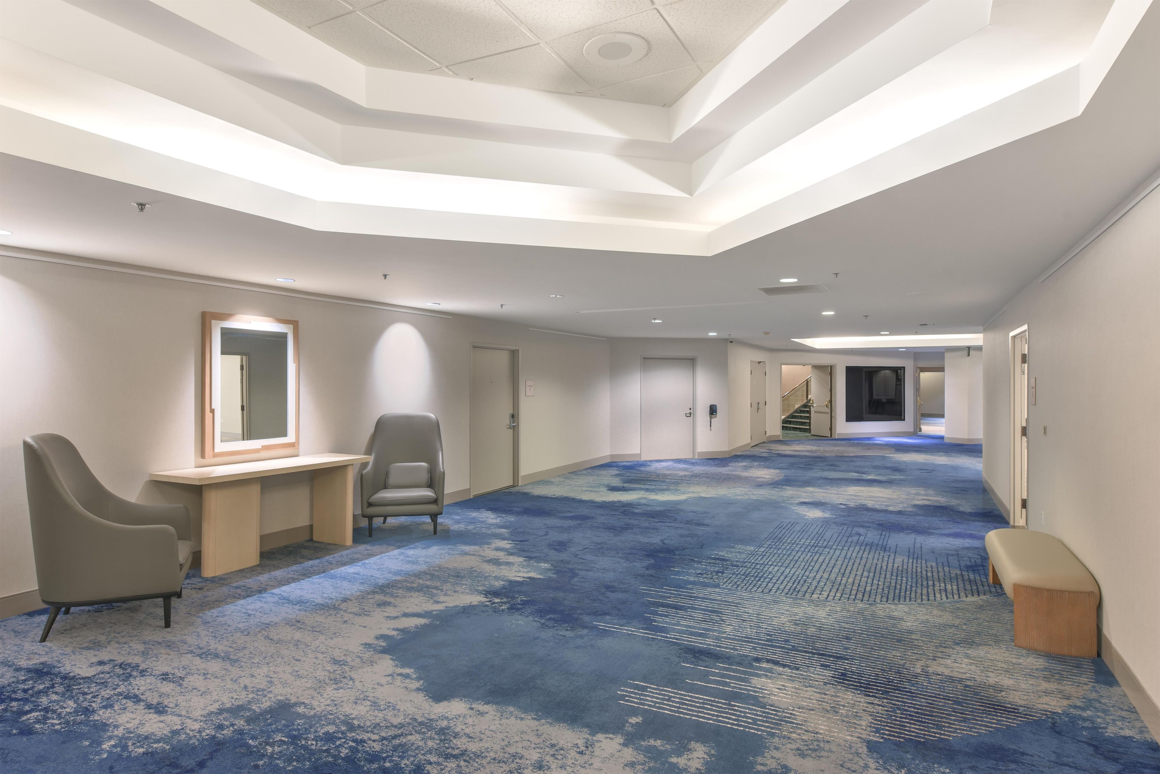 A hotel corridor with a blue carpet, a desk with a chair, and a large mirror.