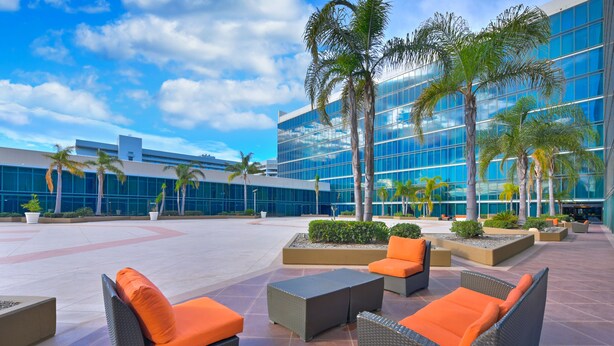 An outdoor seating area with wicker furniture and orange cushions, surrounded by palm trees and a modern building with reflective glass windows.