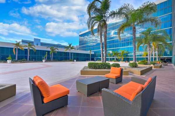An outdoor seating area with wicker furniture and orange cushions, surrounded by palm trees and a modern building with reflective glass windows. - Hilton Anaheim (Anaheim)
