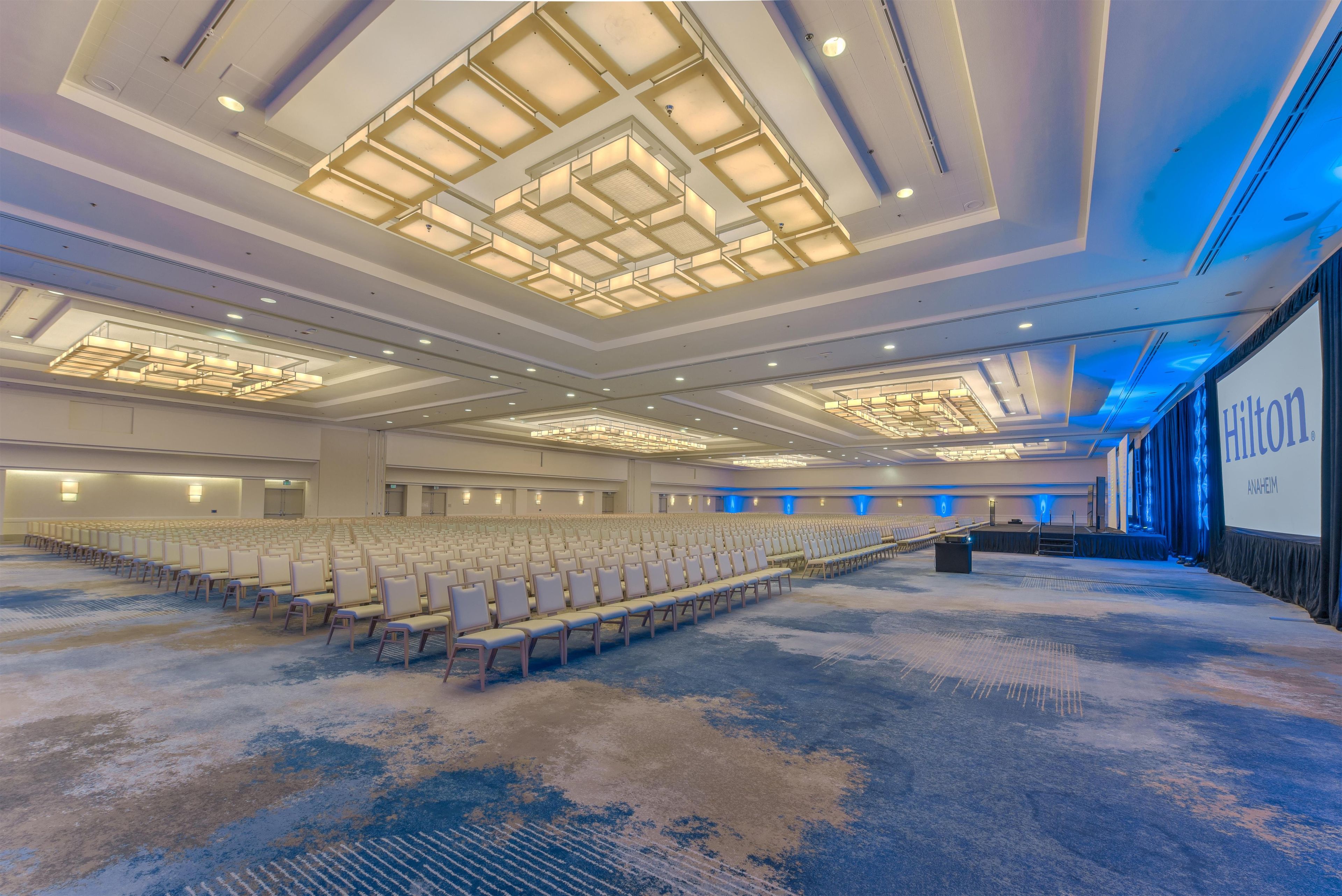 A large conference room with rows of chairs, a stage, and a large screen displaying the Hilton logo.