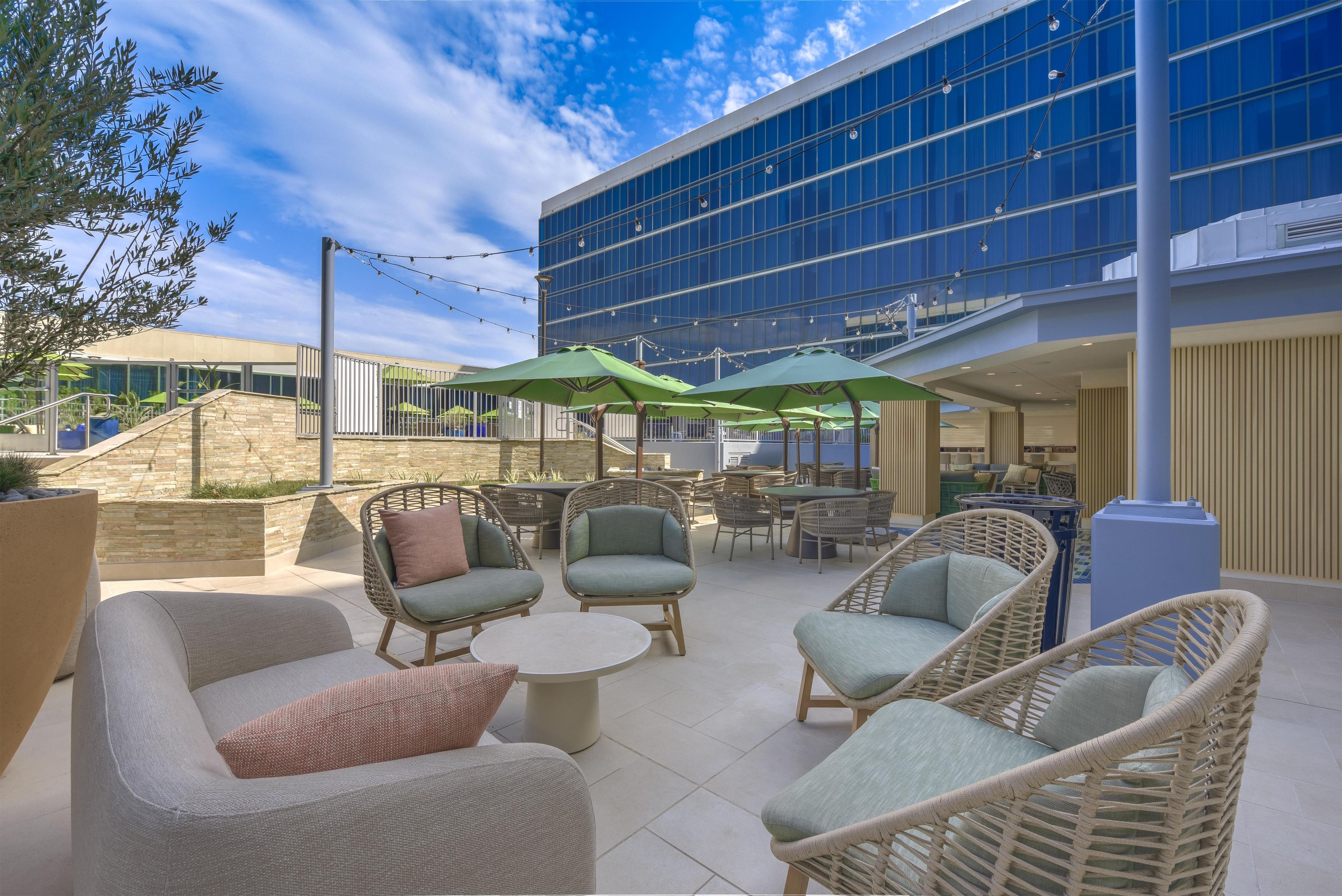 An outdoor seating area with wicker furniture and green umbrellas, a stone wall, and a modern building with large windows in the background.