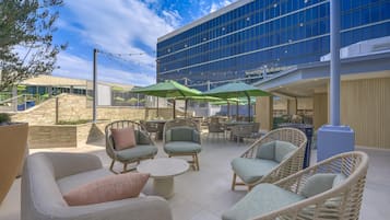 An outdoor seating area with wicker furniture and green umbrellas, a stone wall, and a modern building with large windows in the background.
