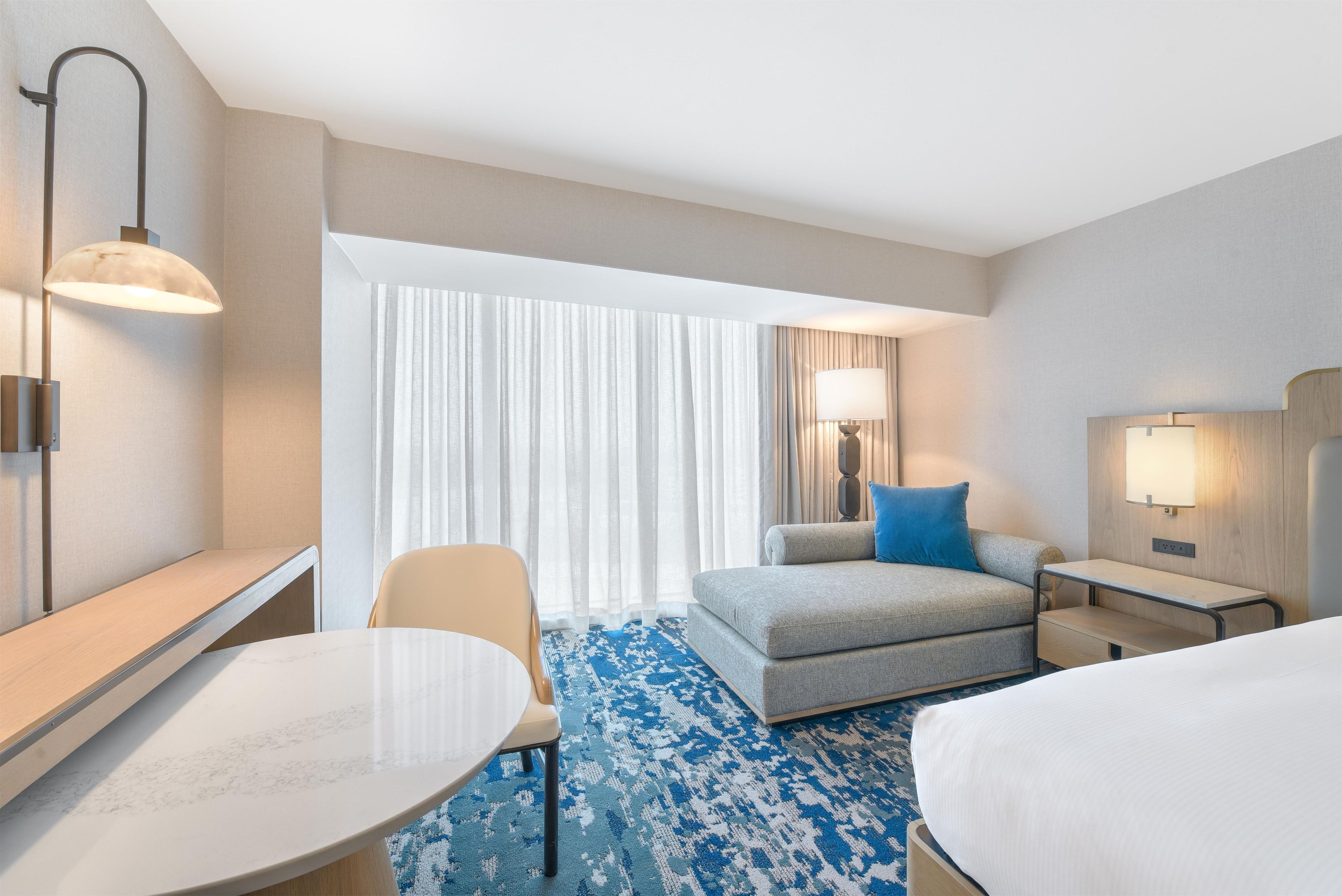 Una habitación de hotel con cama, escritorio, silla, sofá, lámpara y ventana con cortinas.
