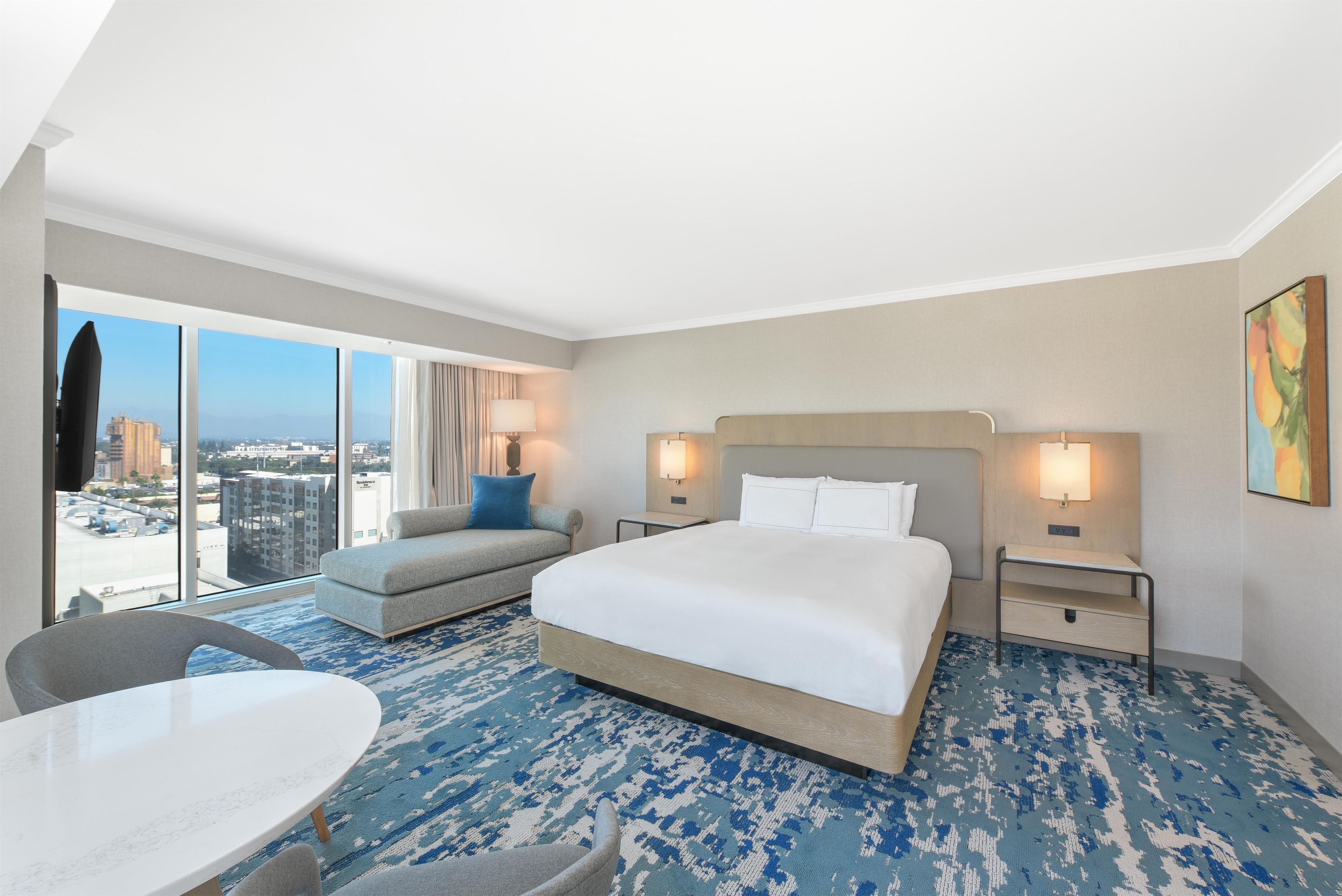 A hotel room with a large bed, a sofa, a dining table, and a view of the city.