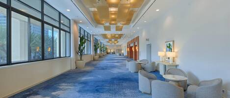 A hotel corridor with a blue carpet, seating area with armchairs, and a view of palm trees outside.