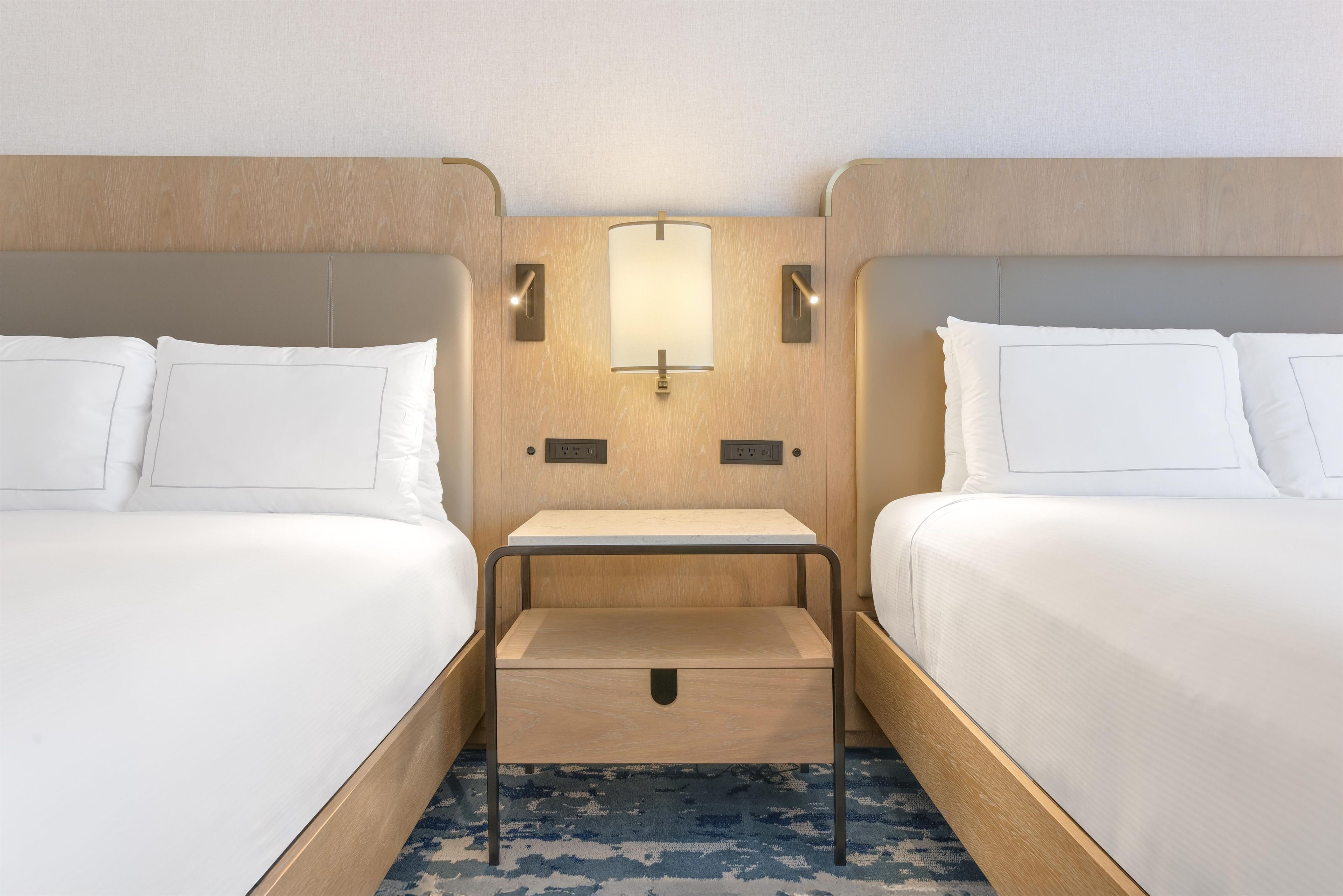 A hotel room with two beds, a bedside table, a lamp, and a power outlet.