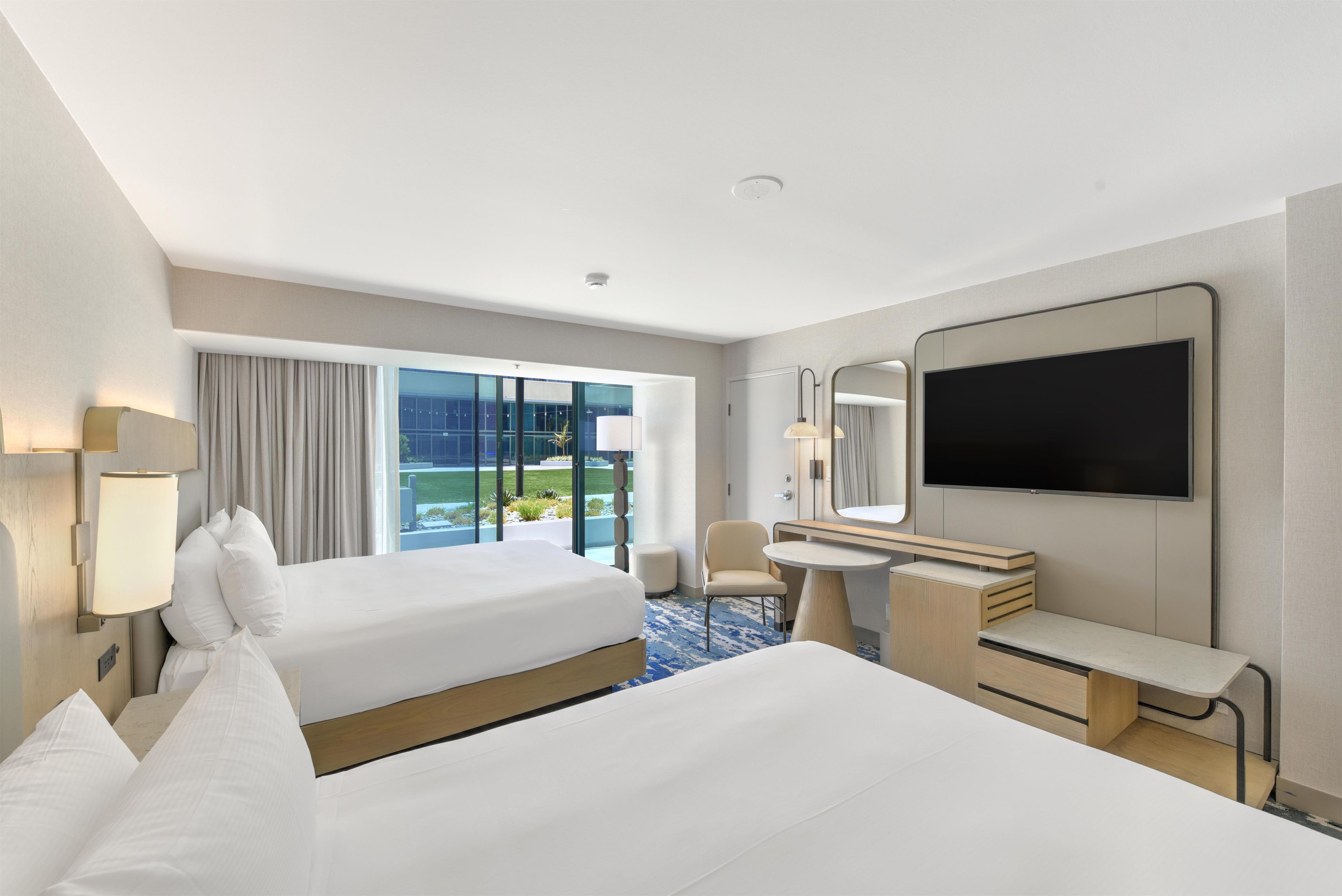 A hotel room with two beds, a desk, a chair, a TV, and a large window offering a view of the outdoors.