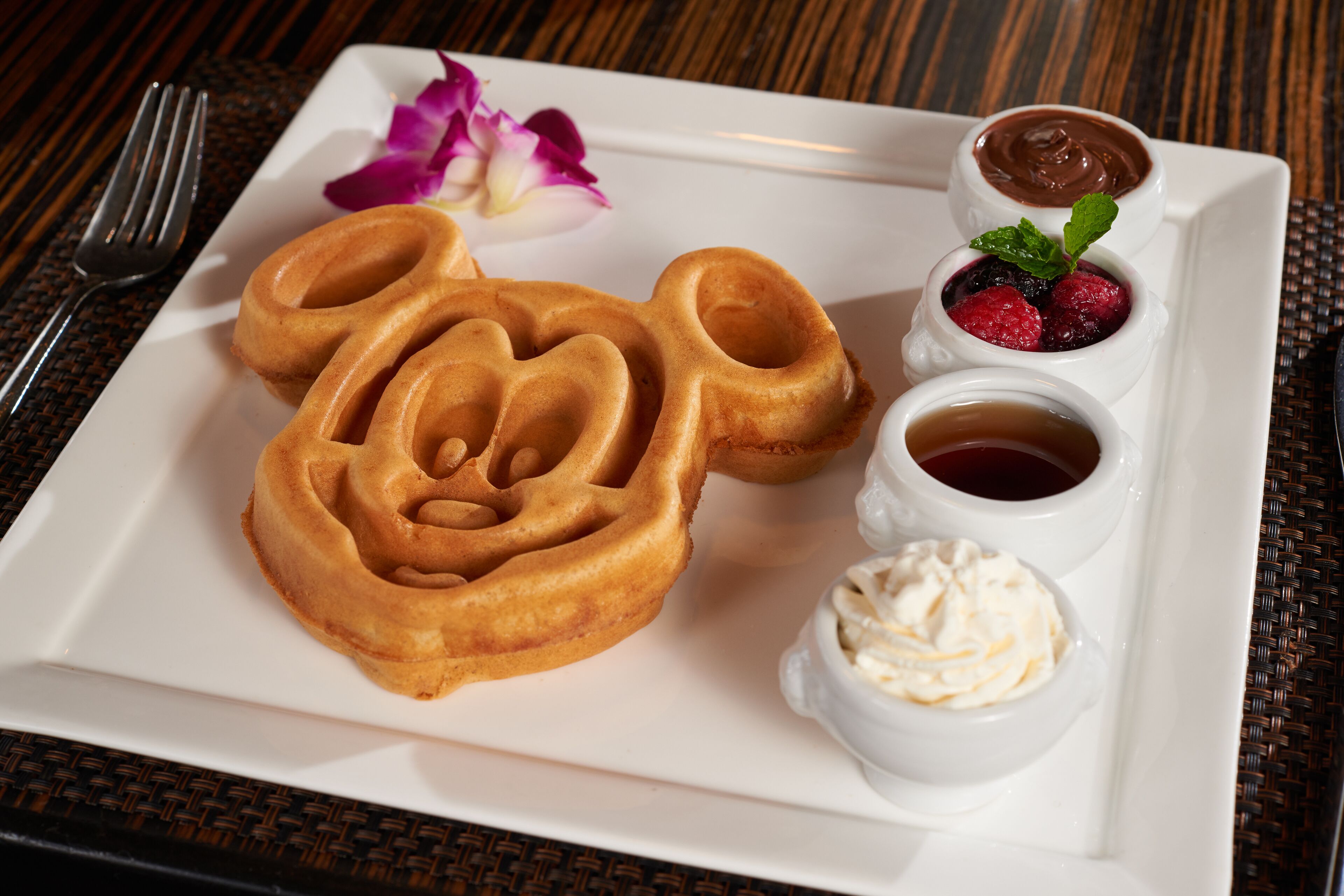 Une assiette avec trois gaufres en forme de Mickey Mouse, un petit bol de sauce au chocolat, un petit bol de fruits rouges, un petit bol de crème fouettée et un petit bol de sirop.