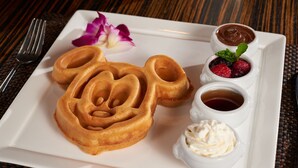 A plate with three waffles shaped like Mickey Mouse, a small bowl of chocolate sauce, a small bowl of berries, a small bowl of whipped cream, and a small bowl of syrup.