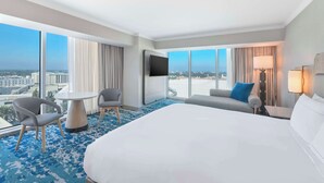 A hotel room with a large bed, a TV, a small table, two chairs, a sofa, and a view of the city.