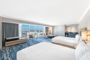Habitación de hotel con dos camas, un escritorio, una silla, una mesita, un sofá, zona de cocina y grandes ventanales con vista a la ciudad.