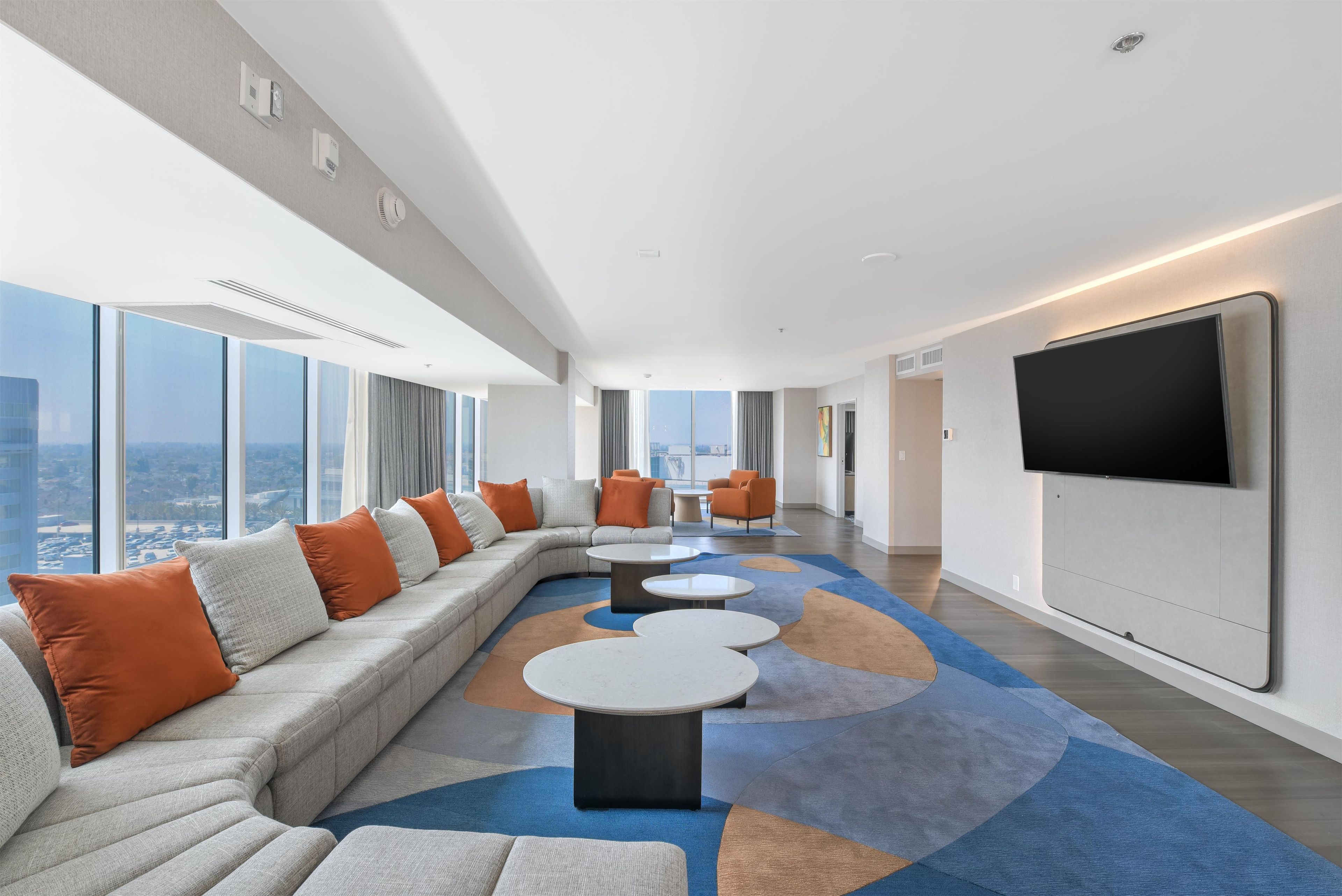 A modern hotel room with a large sectional sofa, a flat-screen TV mounted on the wall, and a city view through large windows.