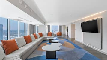 A modern hotel room with a large sectional sofa, a flat-screen TV mounted on the wall, and a city view through large windows.